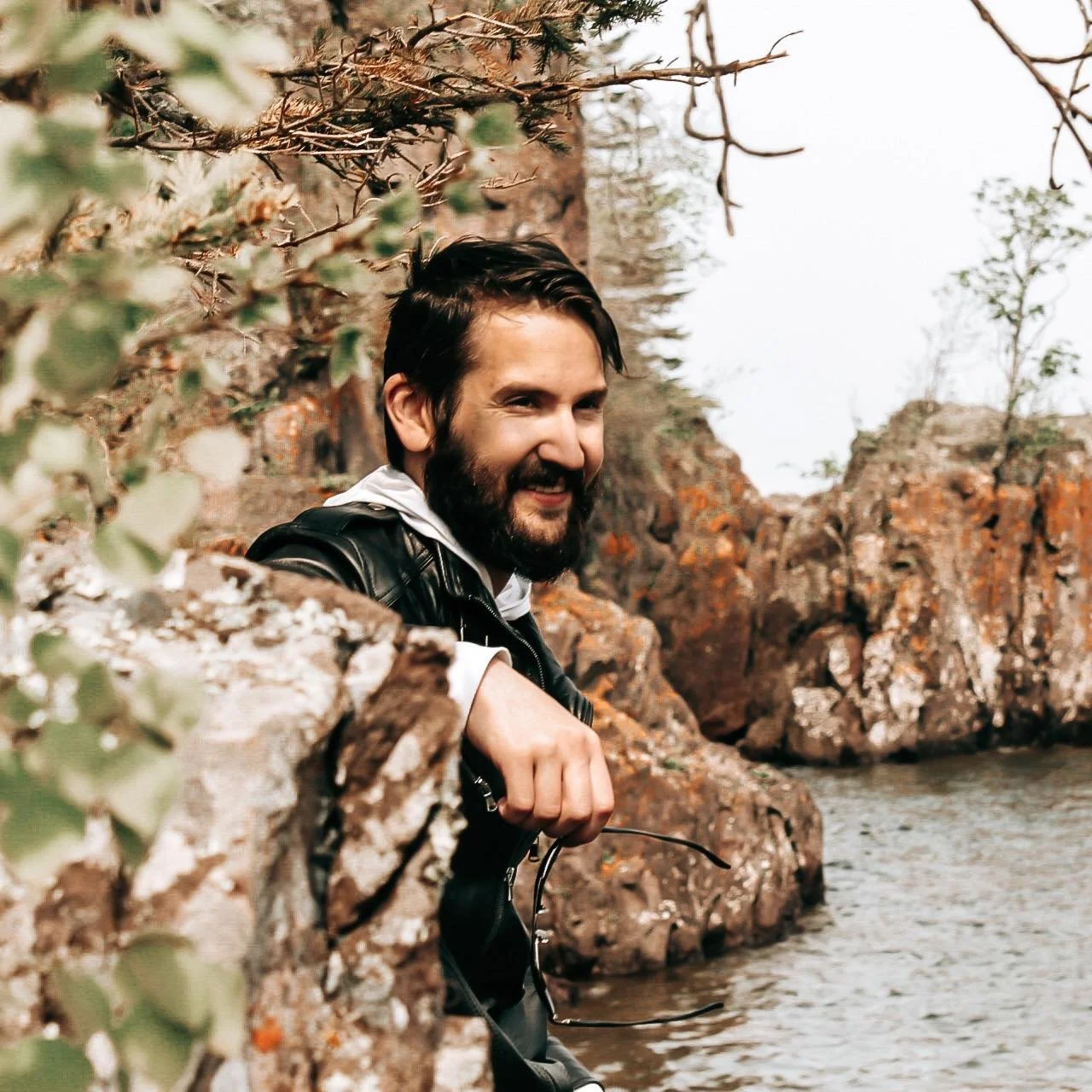 A man with dark hair and a beard smiling while standing by a rocky shoreline surrounded by trees.