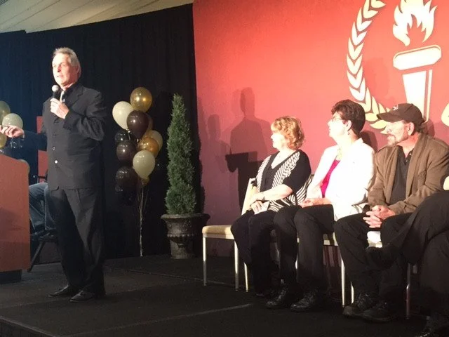 A man in a dark suit speaking into a microphone on stage at a panel event, with three seated women and a man listening. The background has balloons and a large emblem with a torch and wreath.