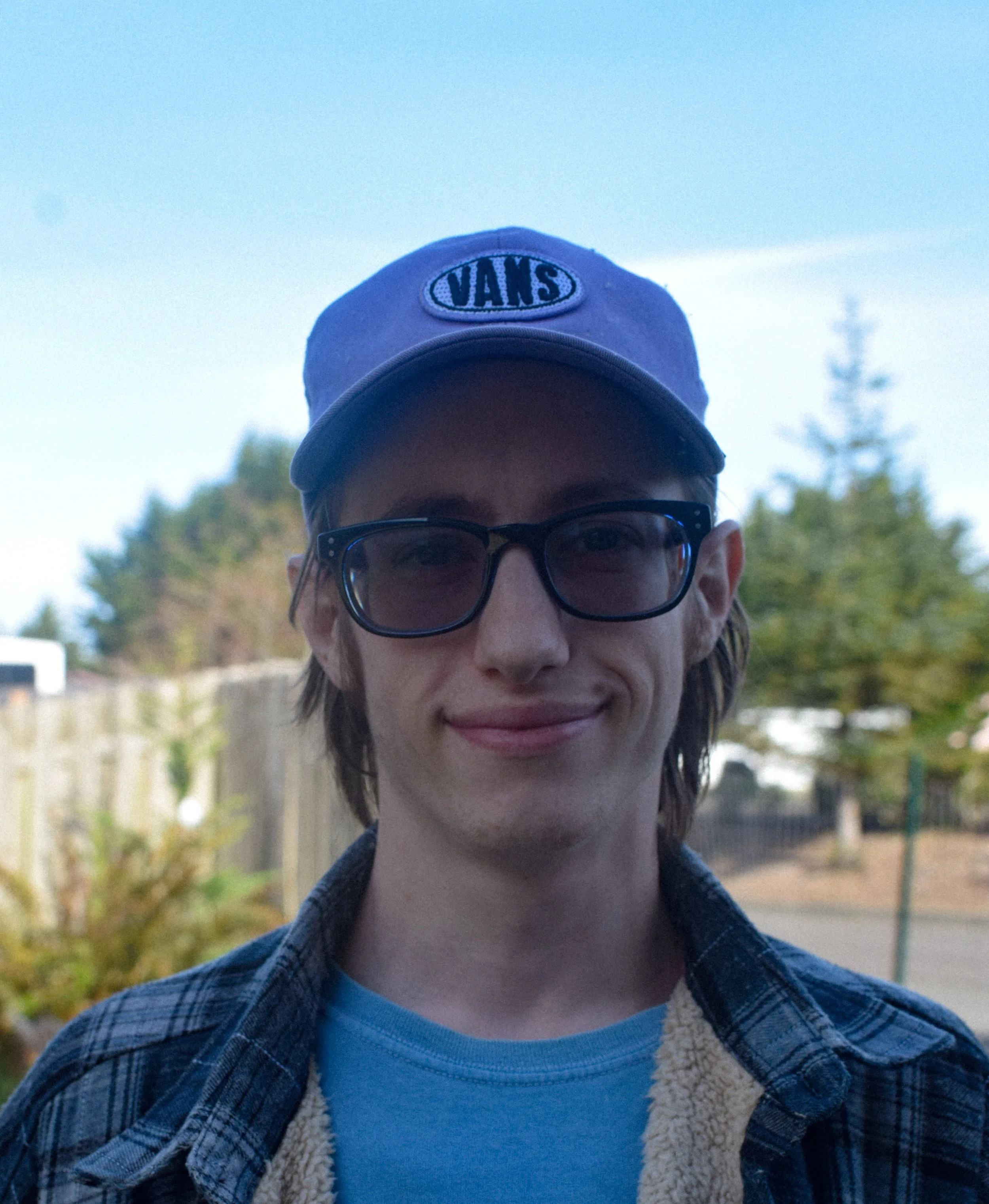 A young man with glasses and a blue cap with the Vans logo, smiling outdoors with trees and a fence in the background.