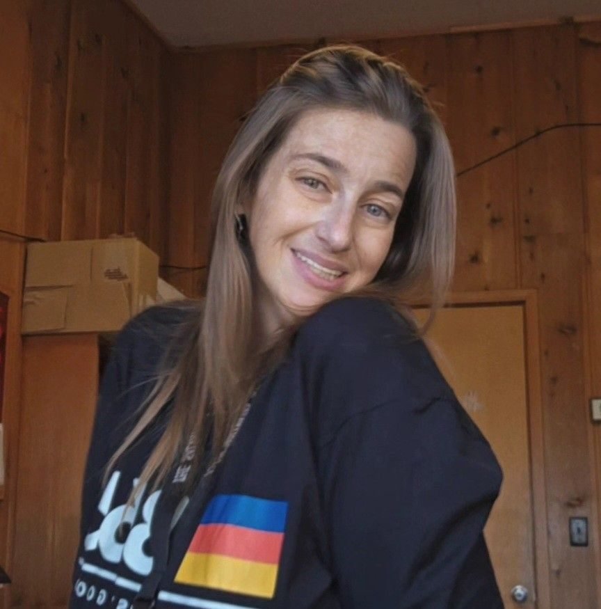 A woman with long brown hair smiling at the camera, wearing a black jacket with a German flag patch on the sleeve, standing in a wooden-paneled room.