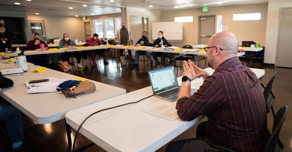 A person at a conference table using a laptop during a meeting with several people seated around a U-shaped arrangement, all wearing masks.