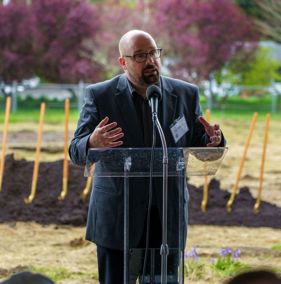 Bald man with glasses, wearing a dark suit, speaking at a podium outdoors with shovels and freshly dug earth in the background, pink flowering tree, and green grass.