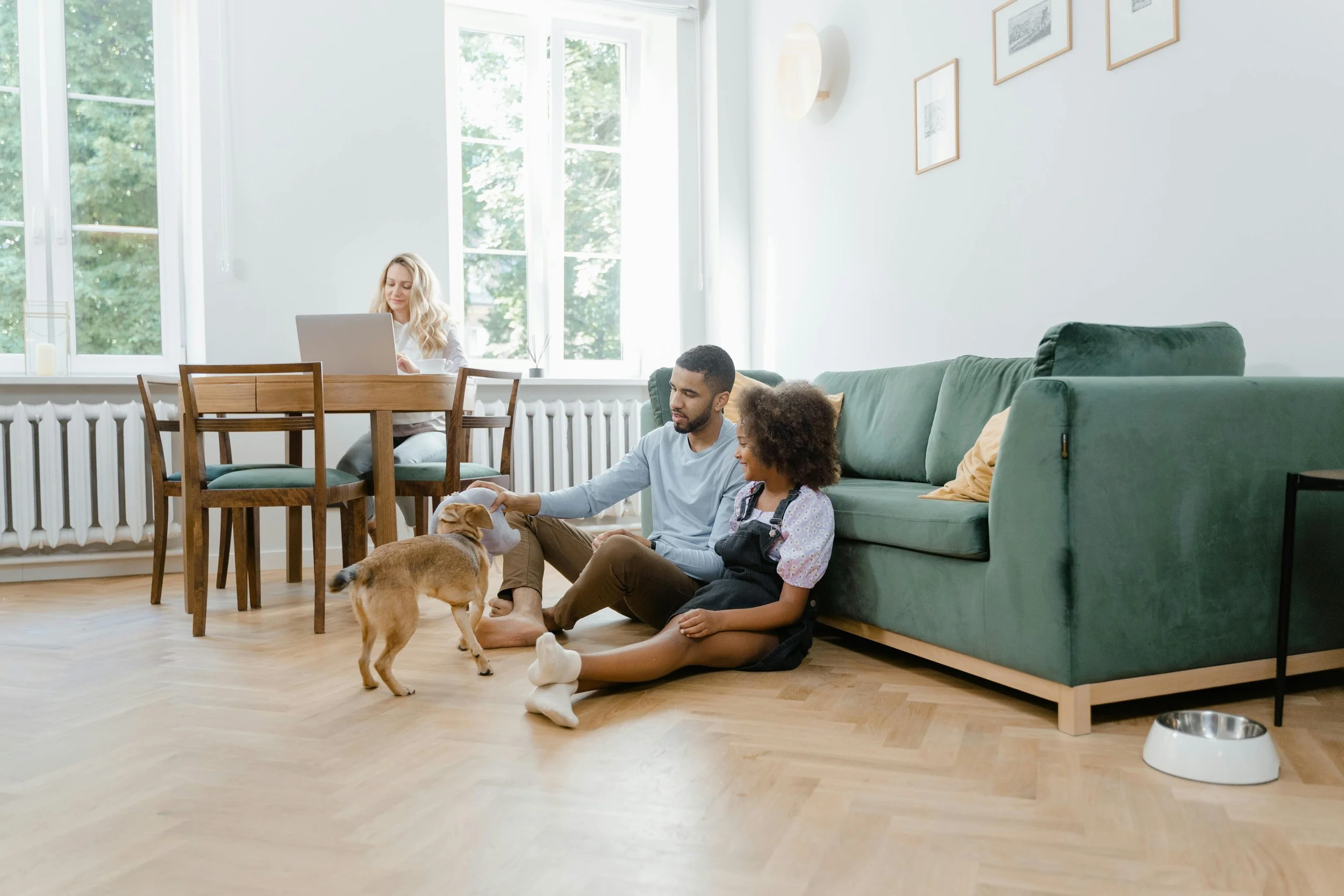 Family indoors with a small dog, sitting on the floor near a green sofa, with a woman working on a laptop at a dining table in a bright room with large windows and framed pictures on the wall.