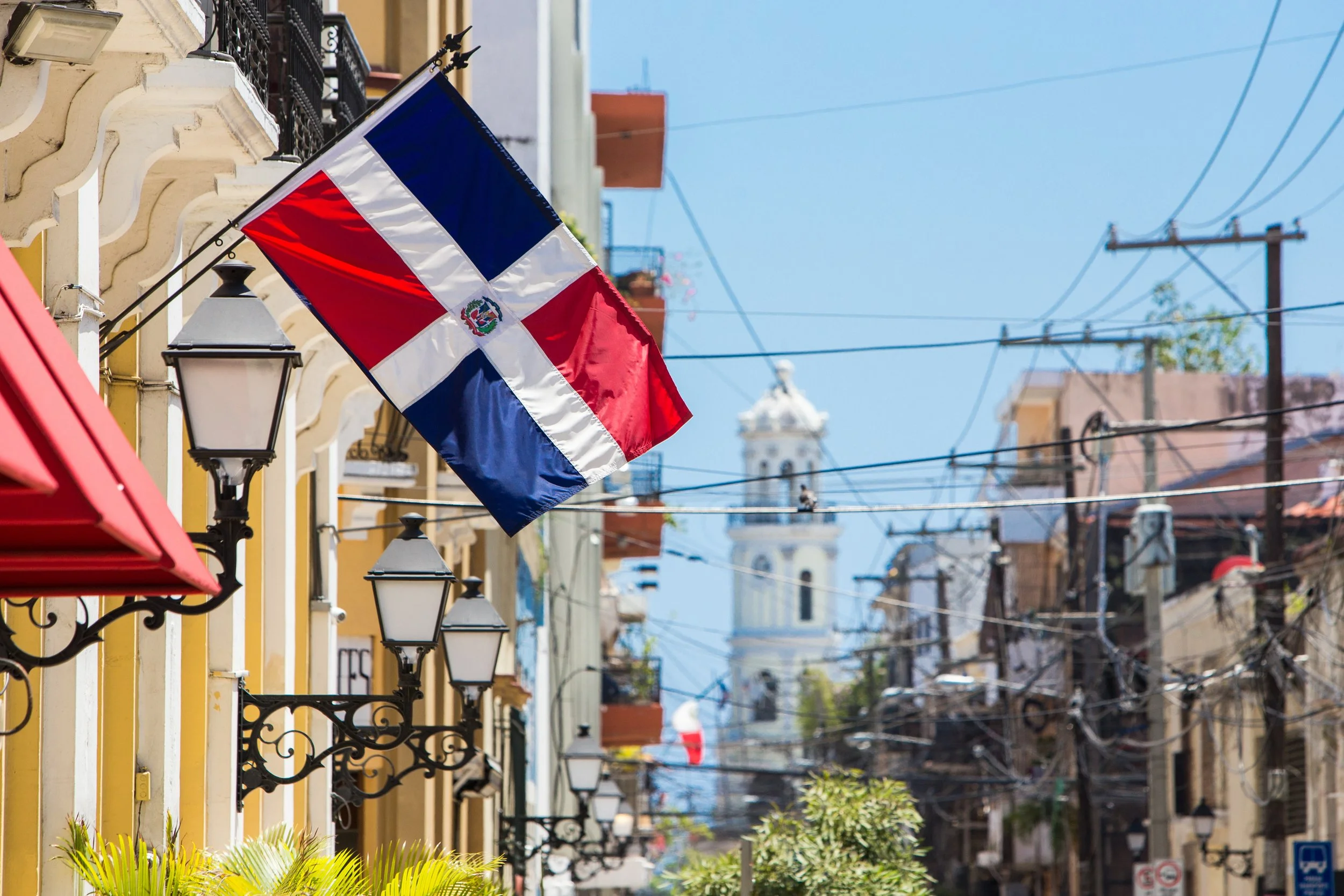 A street scene in a Latin American city with a Dominican Republic flag hanging from a building, colorful buildings, and a clock tower in the background.