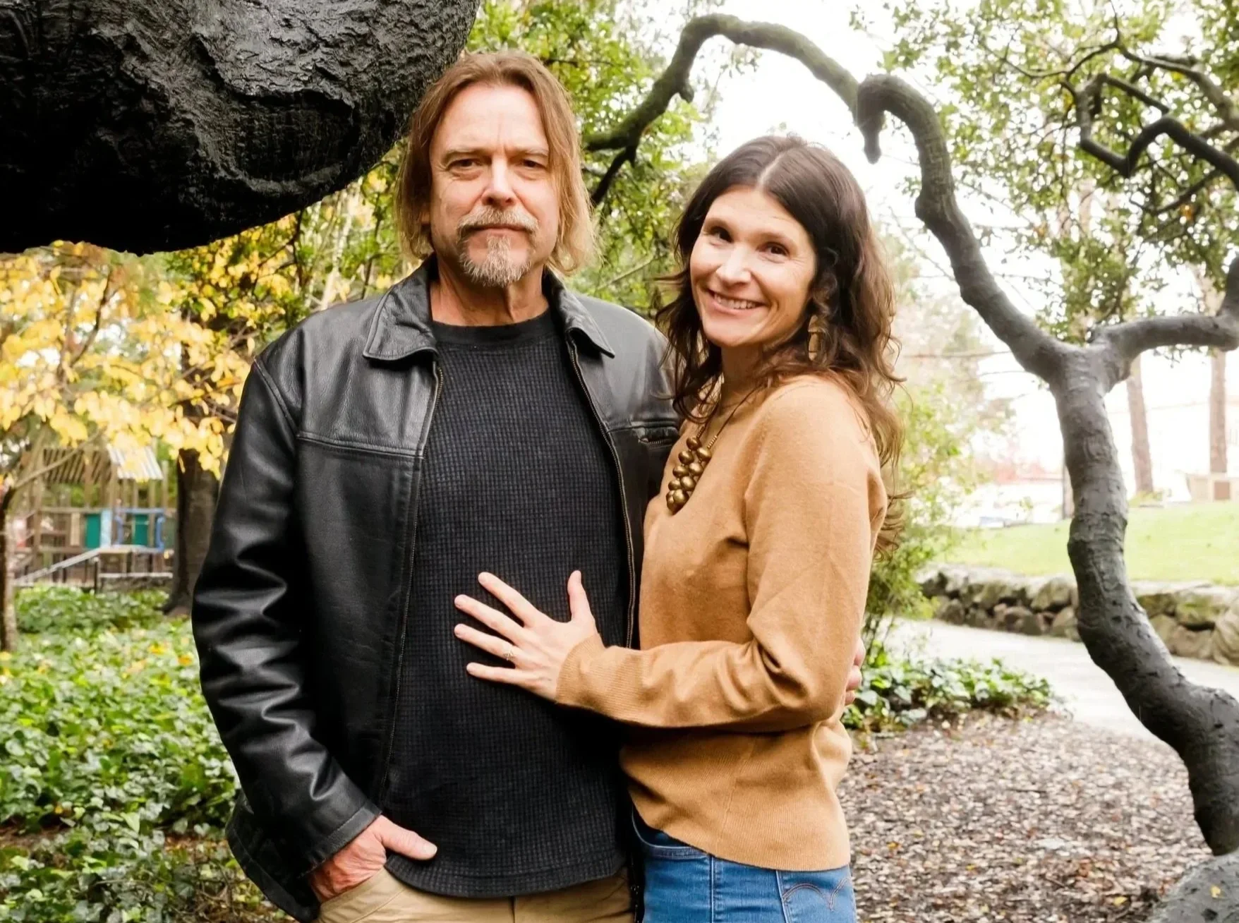 A man and woman standing together outdoors in a park with trees and walking paths in the background. The woman has her hand on the man's chest and is smiling at the camera. The man has a serious expression and is wearing a black leather jacket and black shirt. The woman is wearing a brown sweater and a large necklace.