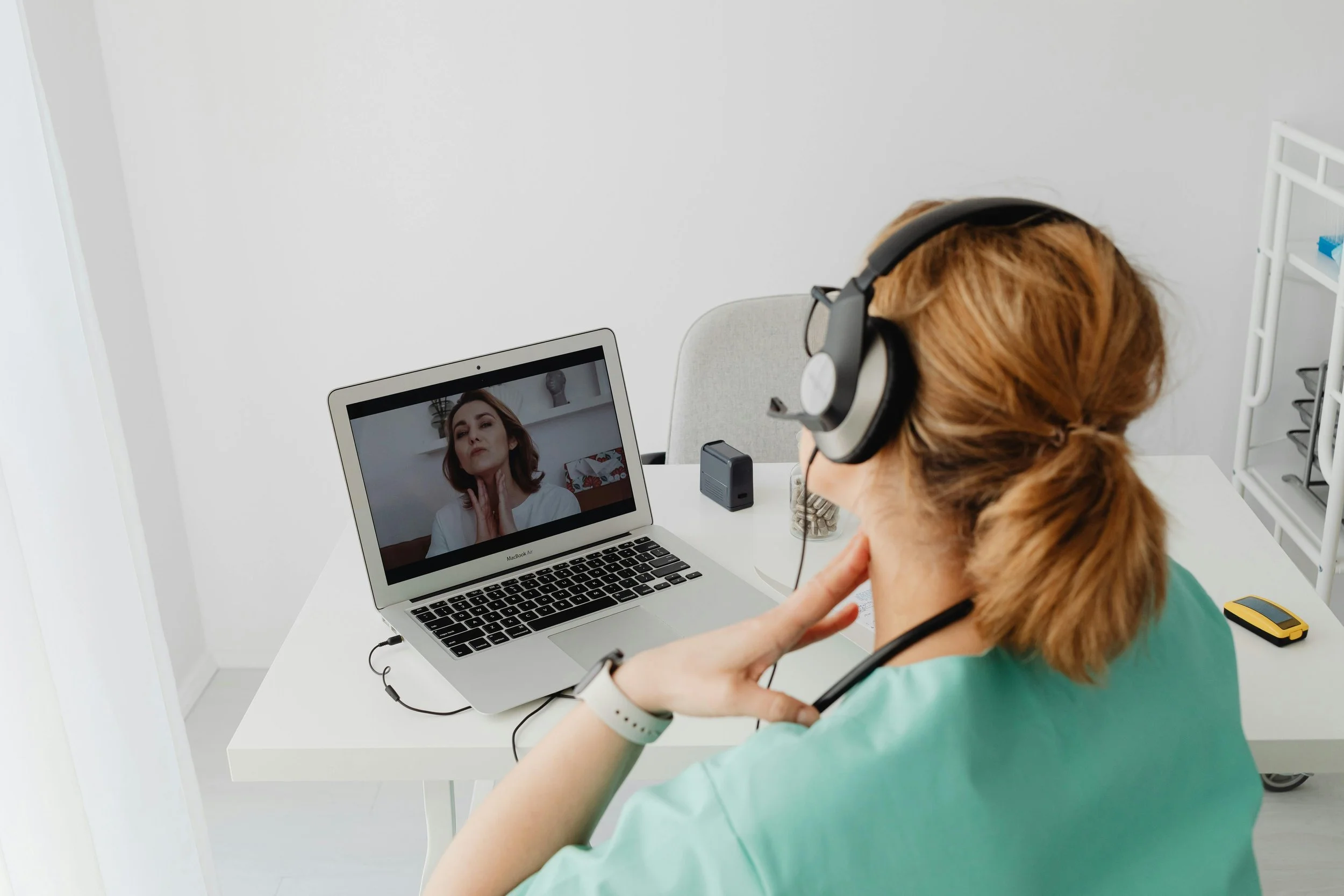 A woman with glasses and a large pair of headphones is sitting at a white desk with a laptop, on a video call with another woman, who has brown hair and is touching her neck.