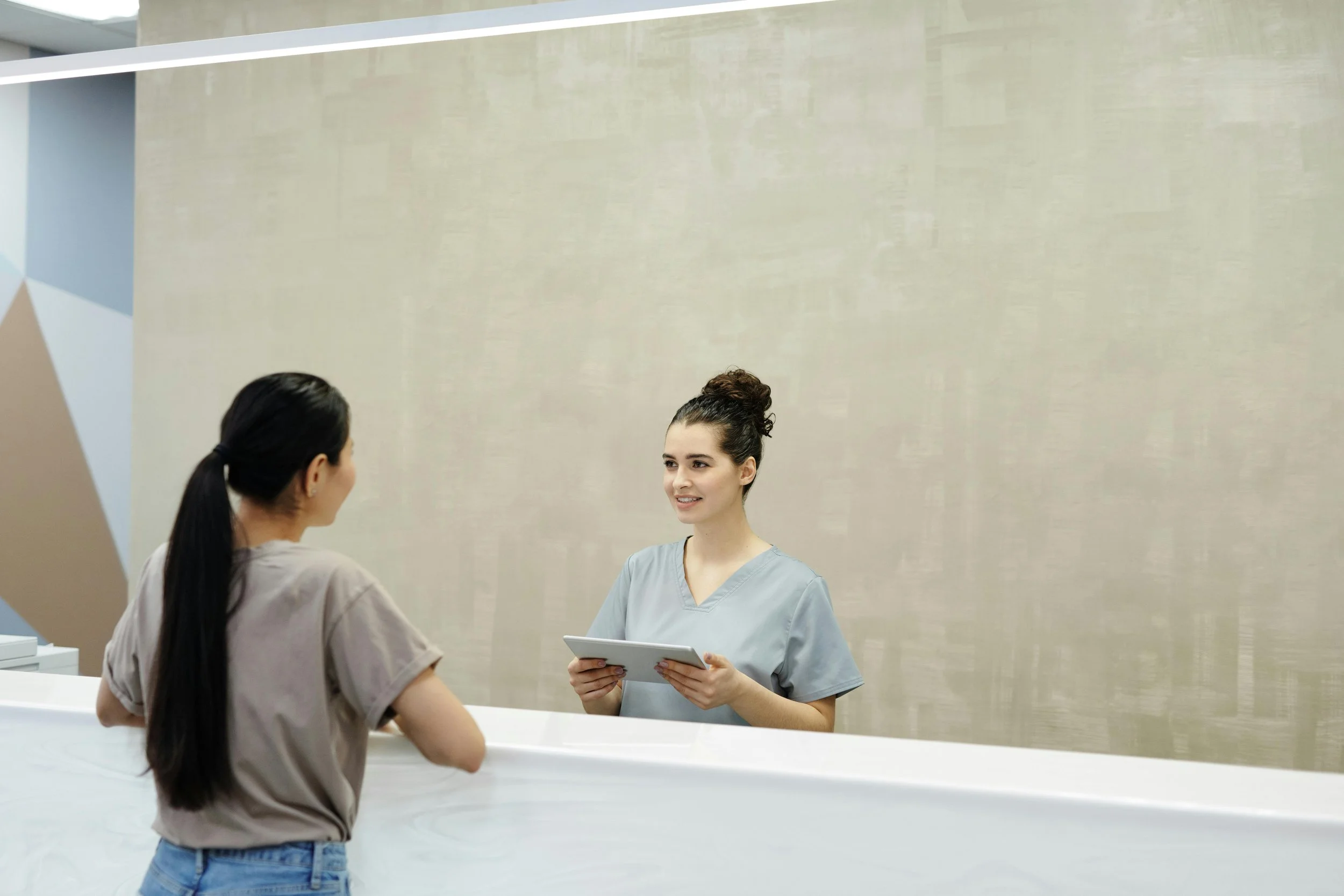 A woman checking in at a reception desk in a modern medical or dental office while smiling and talking to a receptionist on the other side of the counter.