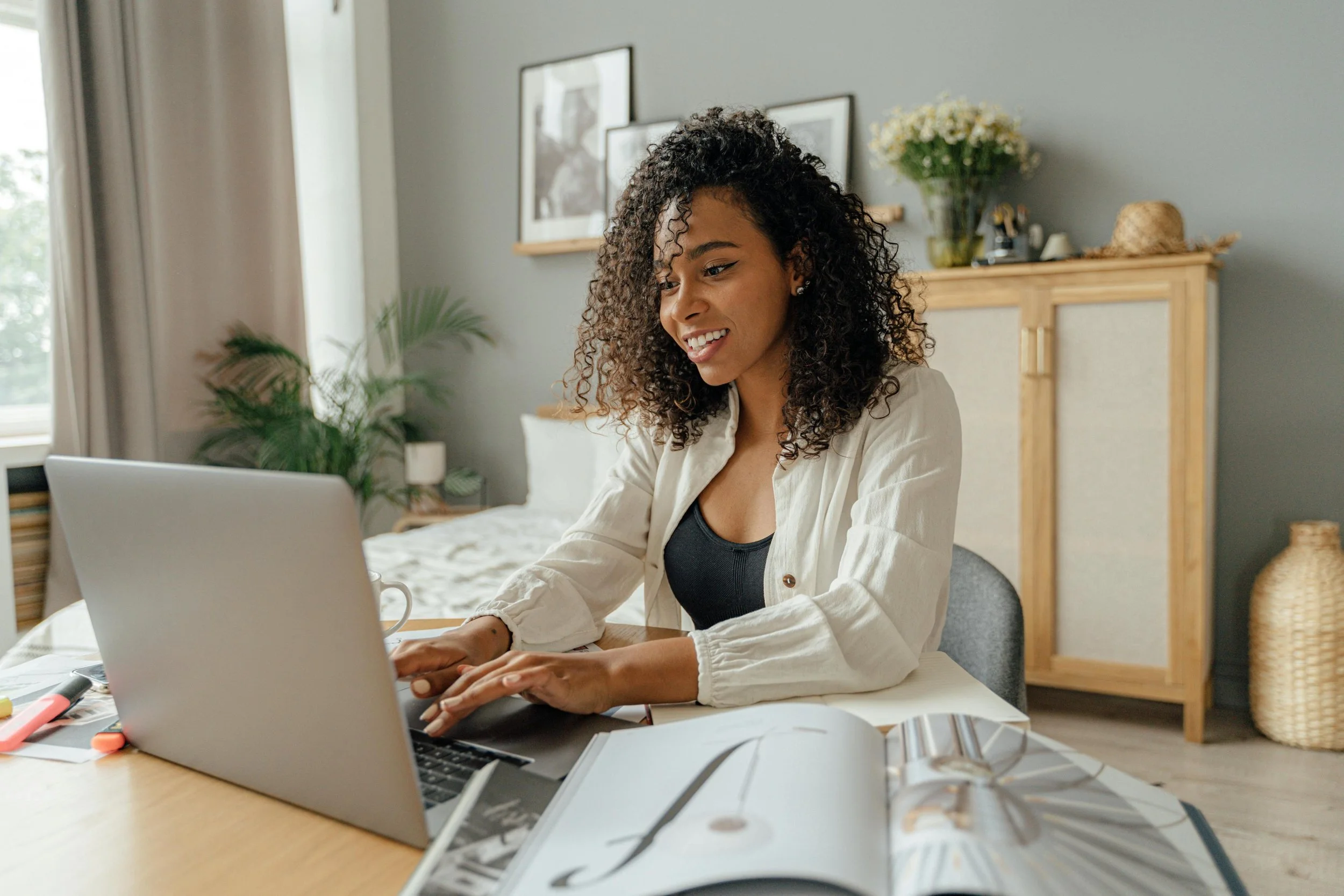 A woman with curly hair smiling while working on a laptop at a desk in a cozy bedroom.