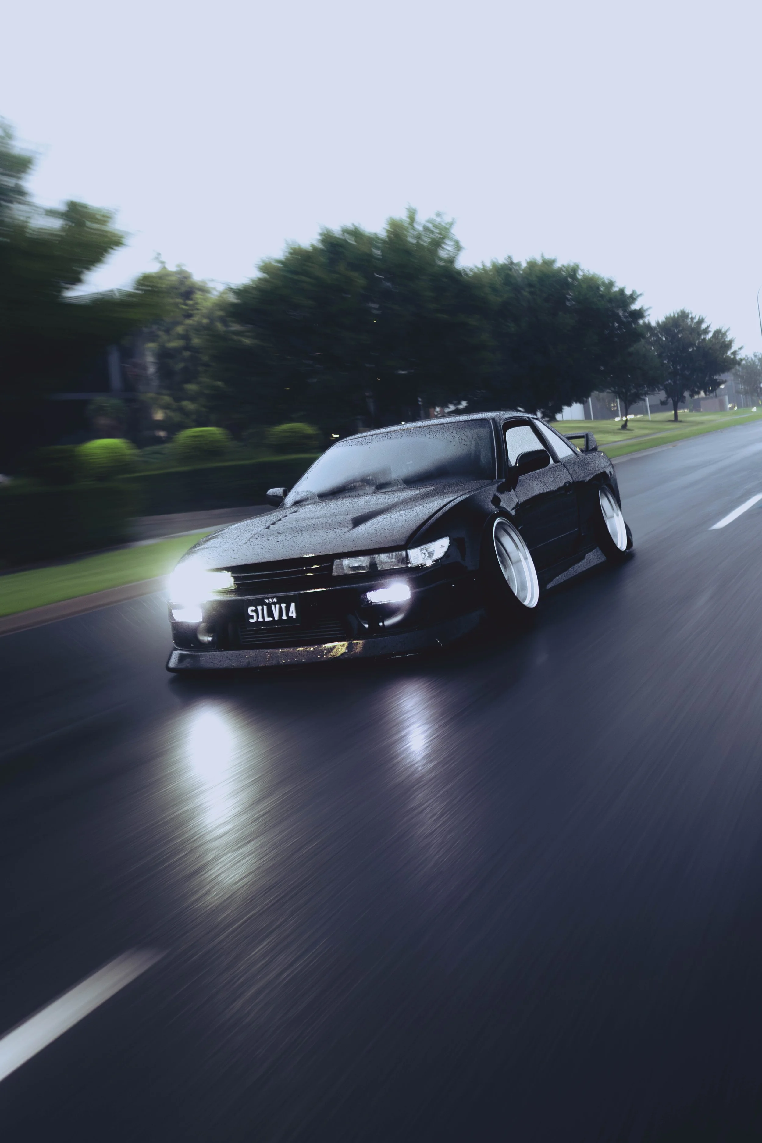 A Stanced Nissan S13 Silvia 240sx with lowered suspension driving on a wet road on a rainy day.