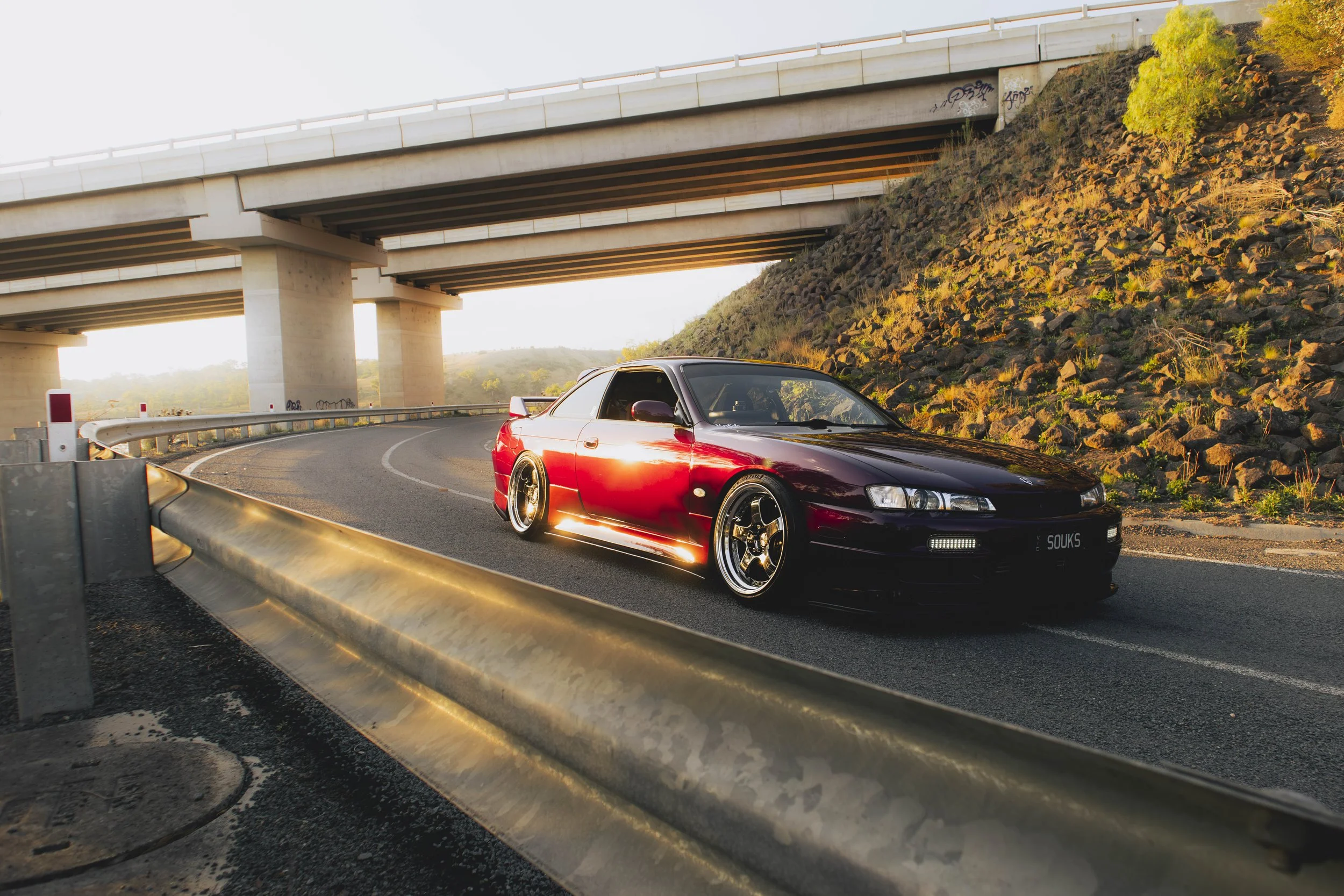 A red sports car with black accents driving on a curved road under an overpass during sunset.