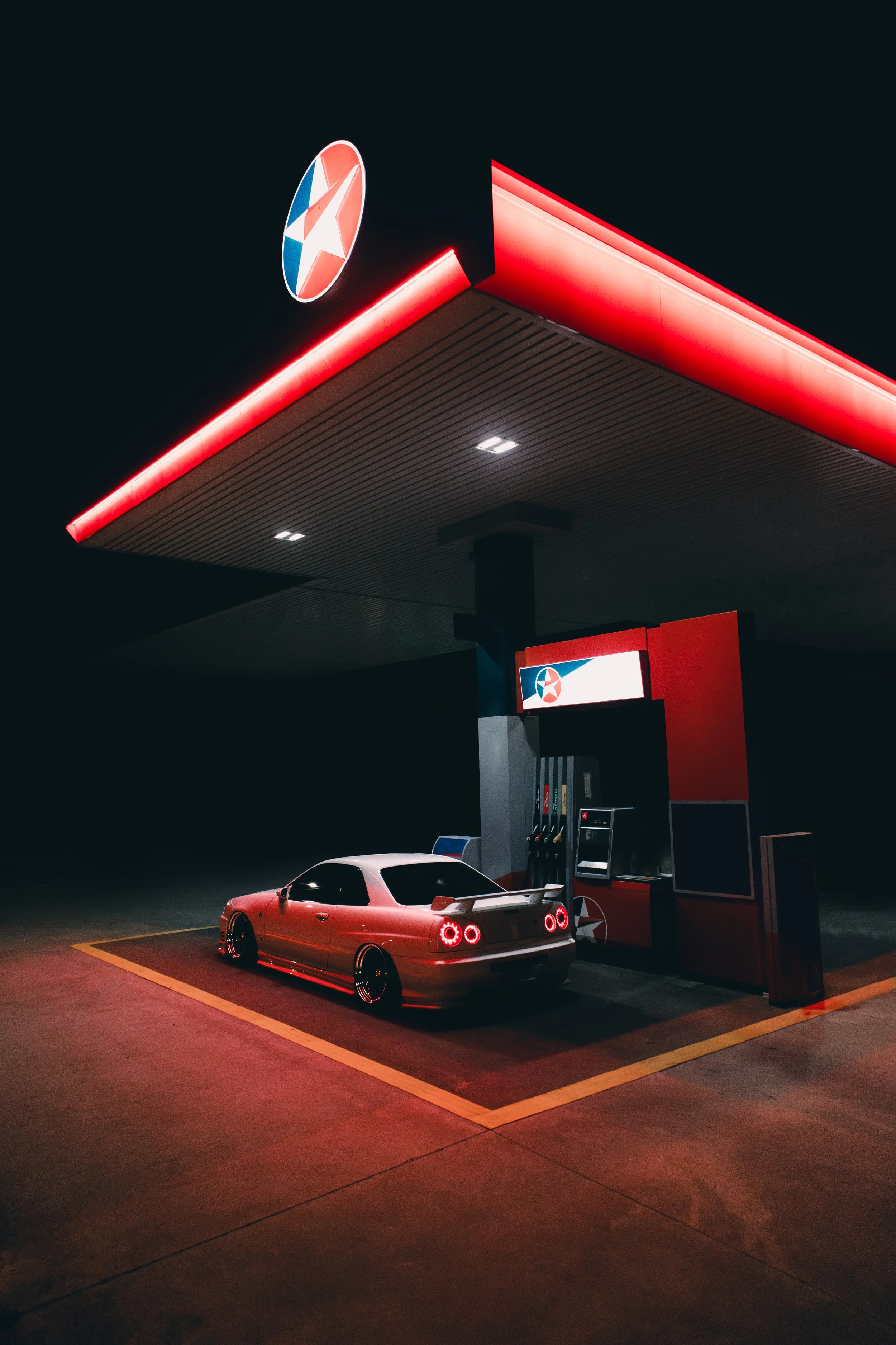 A Nissan Skyline R34 parked at a gasoline station during nighttime, illuminated by red lights, with a gas pump and a bright sign.