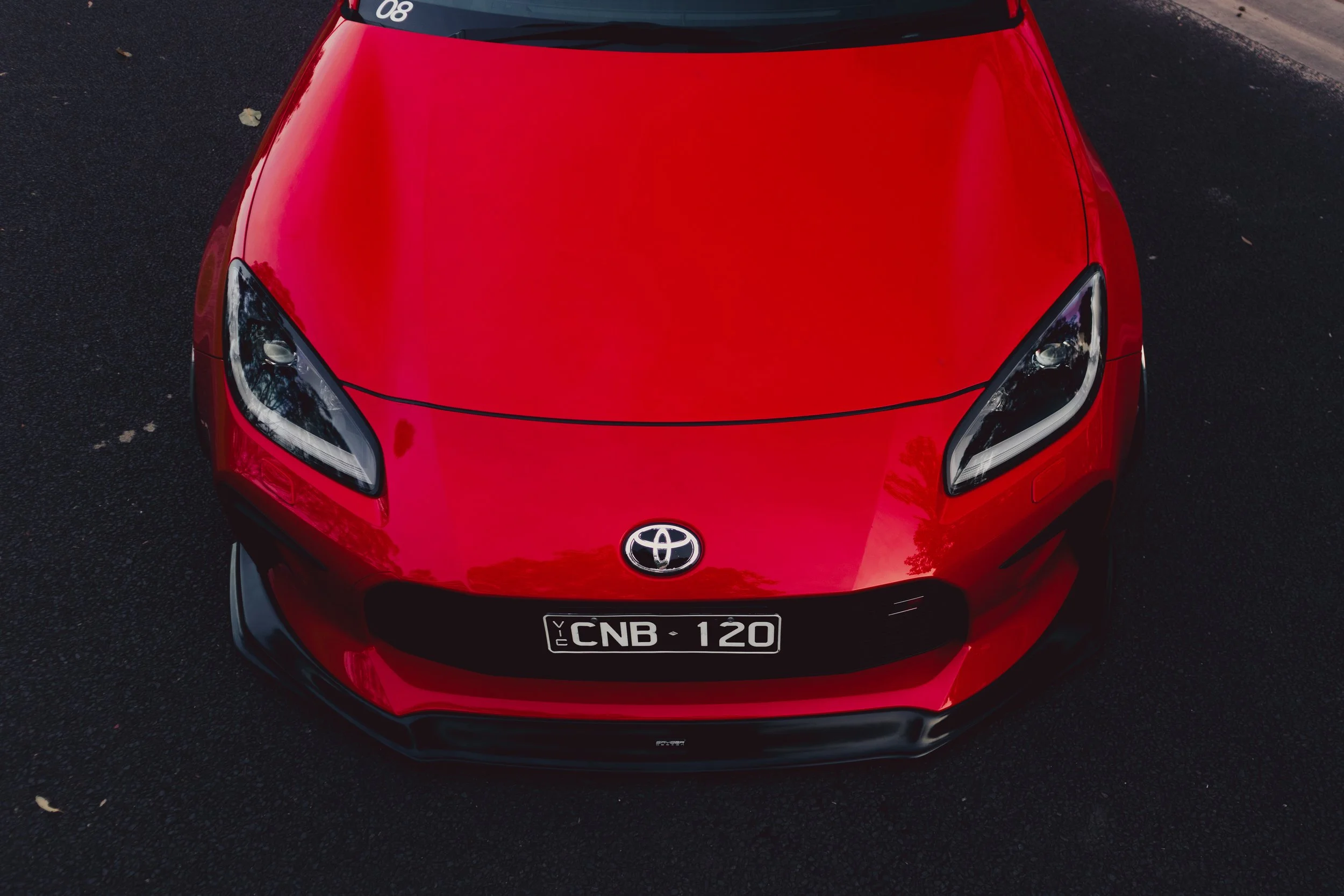 A top view of a red Toyota car parked on a dark asphalt surface, showing the front end, headlights, and license plate with the number CNB 120.
