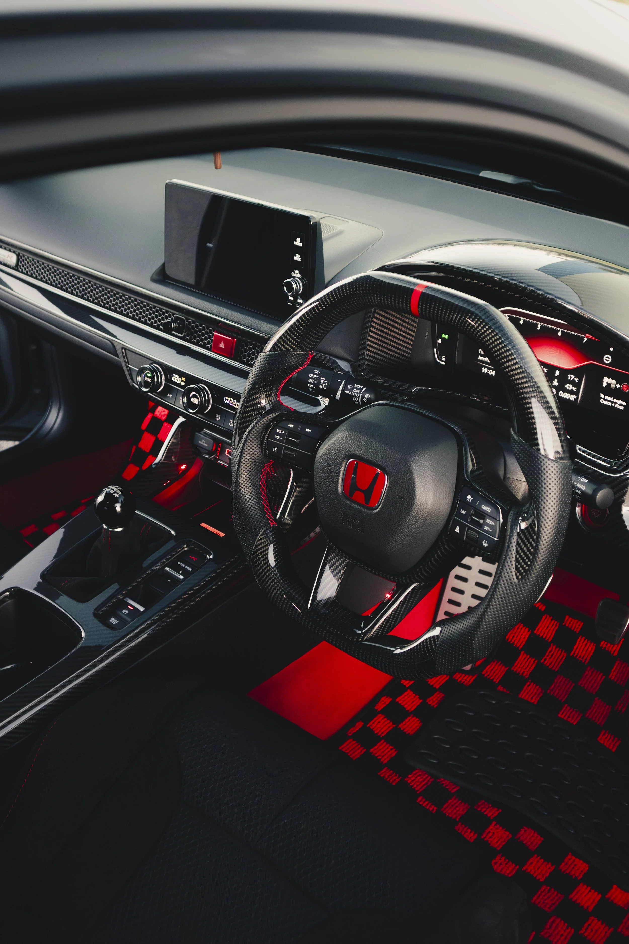 Interior of a sports car with a carbon fiber steering wheel, digital dashboard, and red and black checkered floor mats.