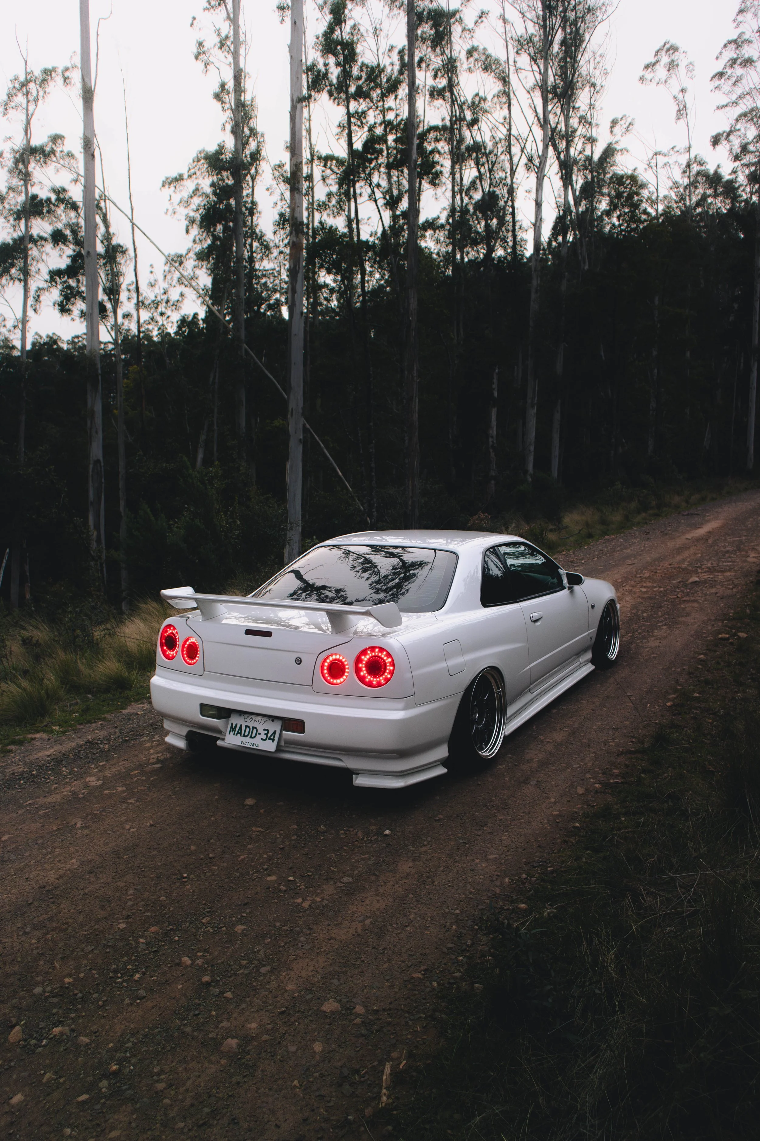 A white sports car with a rear spoiler parked on a dirt road near a wooded area with tall, thin trees.