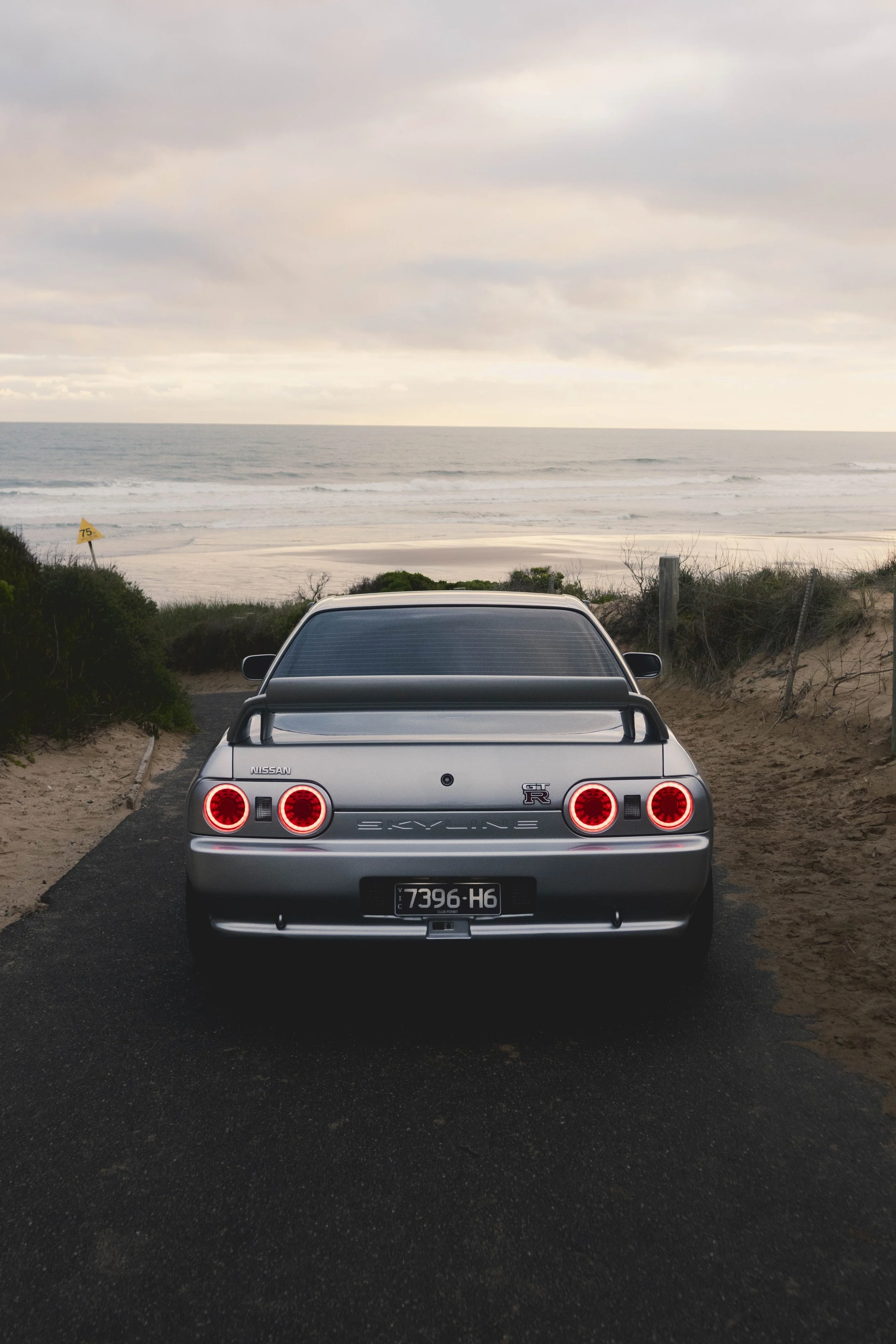 A silver Nissan Skyline R32 GTR car parked on a paved pathway facing the beach, with the ocean and cloudy sky in the background.