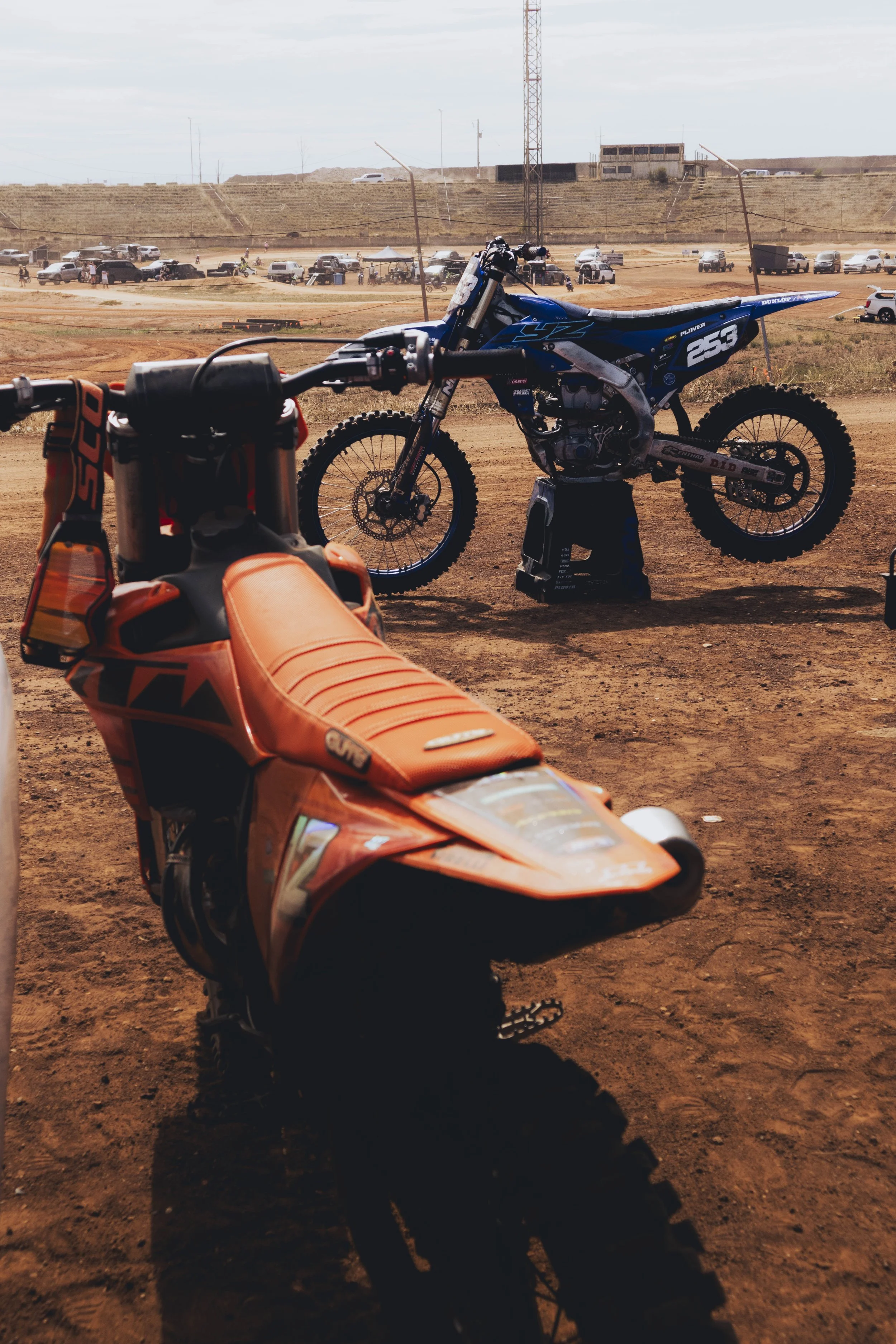 A blue off-road dirt bike on a stand in a dusty motocross track, with a close-up of an orange KTM dirt bike in the foreground, and a background of parked cars and a dirt hill.