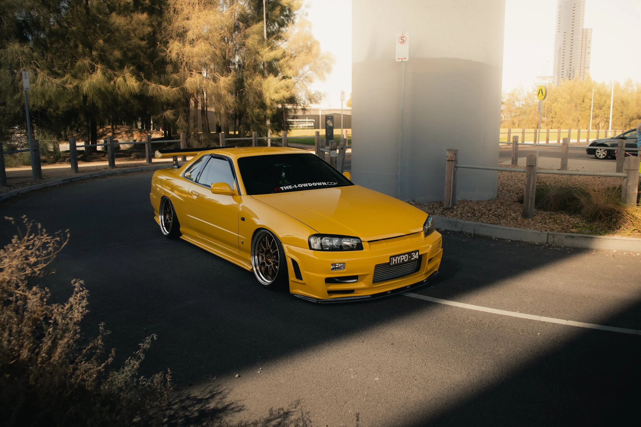 A yellow Nissan Skyline R34 parked in an outdoor lot under a bridge or overpass. The car has a spoiler and aftermarket wheels. 