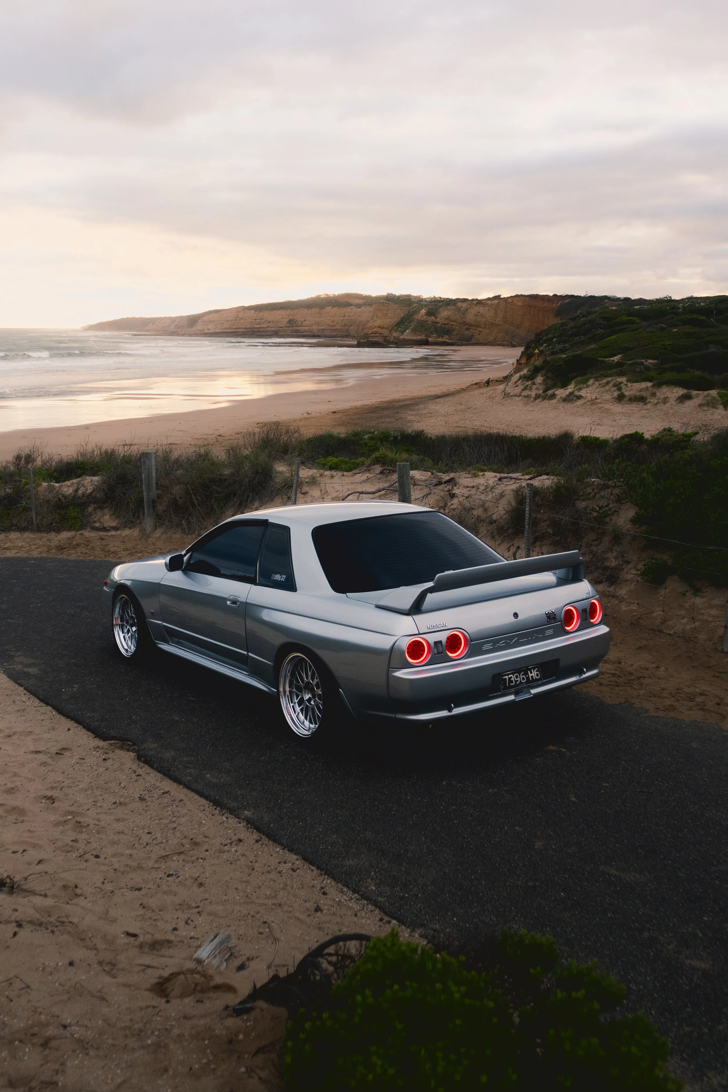 Silver Nissan Skyline R32 GTR parked near a sandy beach with ocean waves and cliffs in the background.