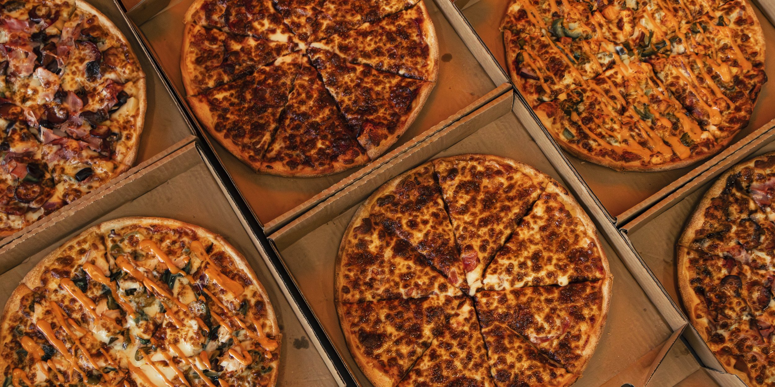 Multiple pizzas in pizza boxes, featuring various toppings including meats, vegetables, and drizzles of sauce.