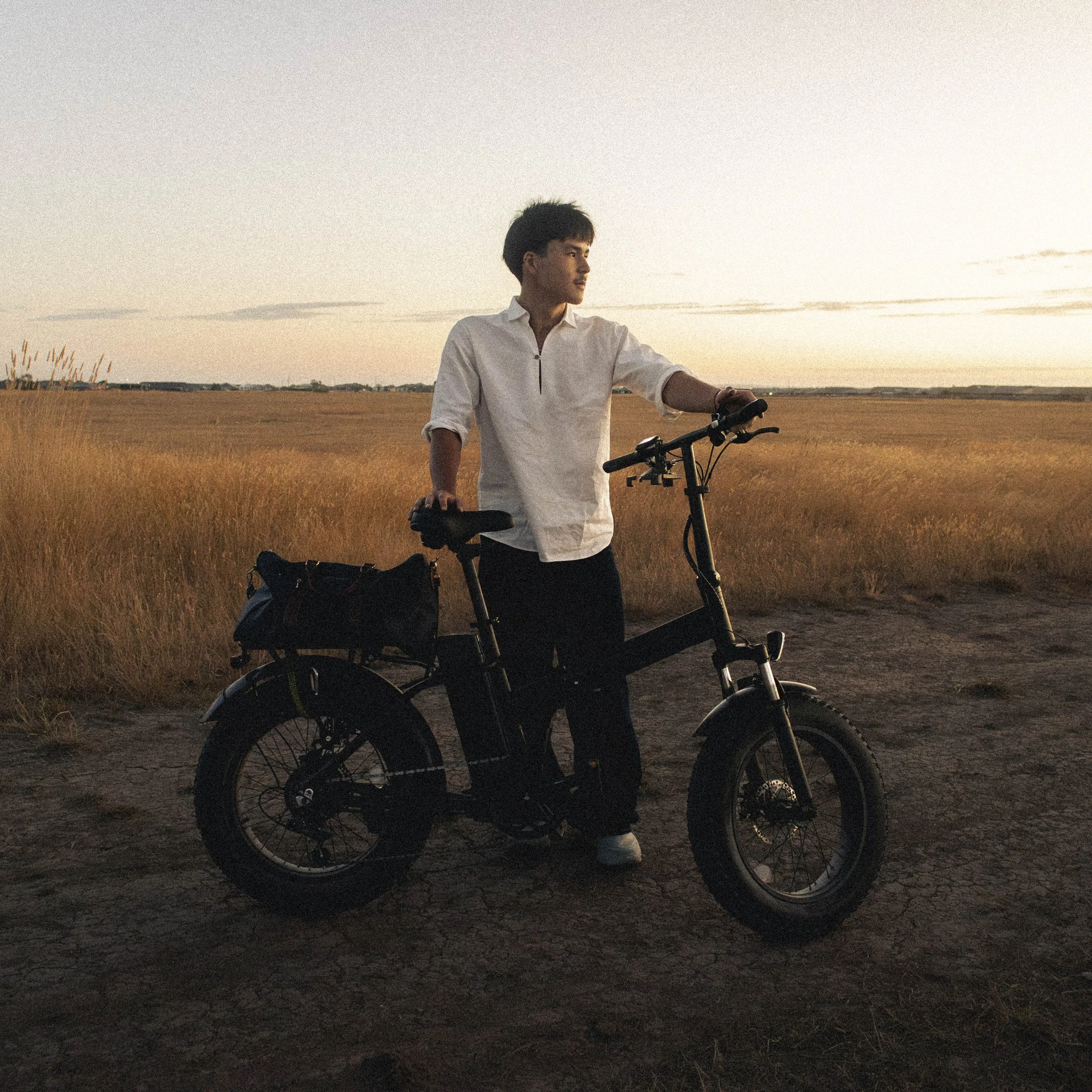 Young man wearing white shirt standing with an electric bicycle in a rural landscape at sunset.