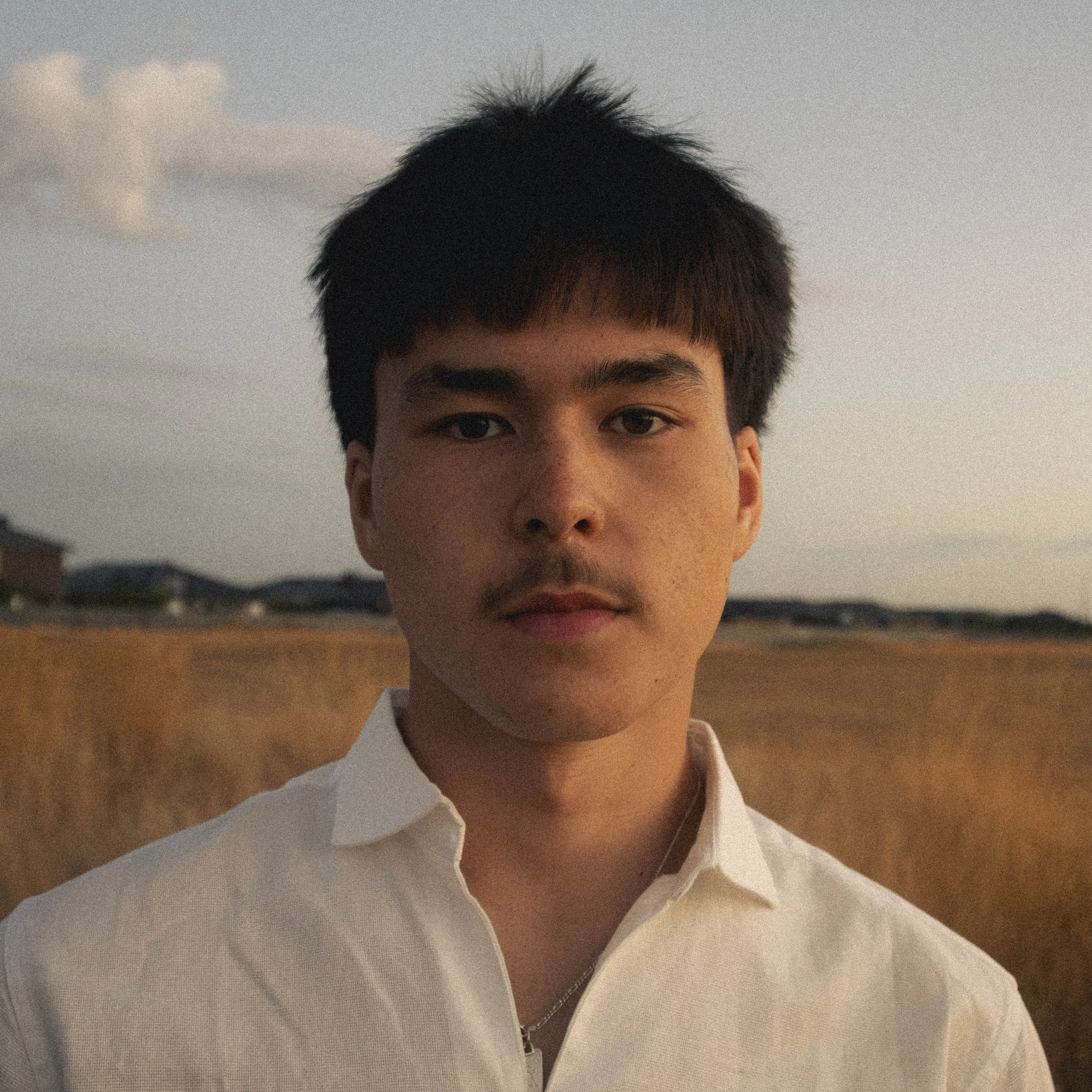 A close-up of a young person with short, dark hair wearing a white collared shirt and a chain necklace, standing outdoors during dusk or sunset with a cloudy sky and hills in the background.