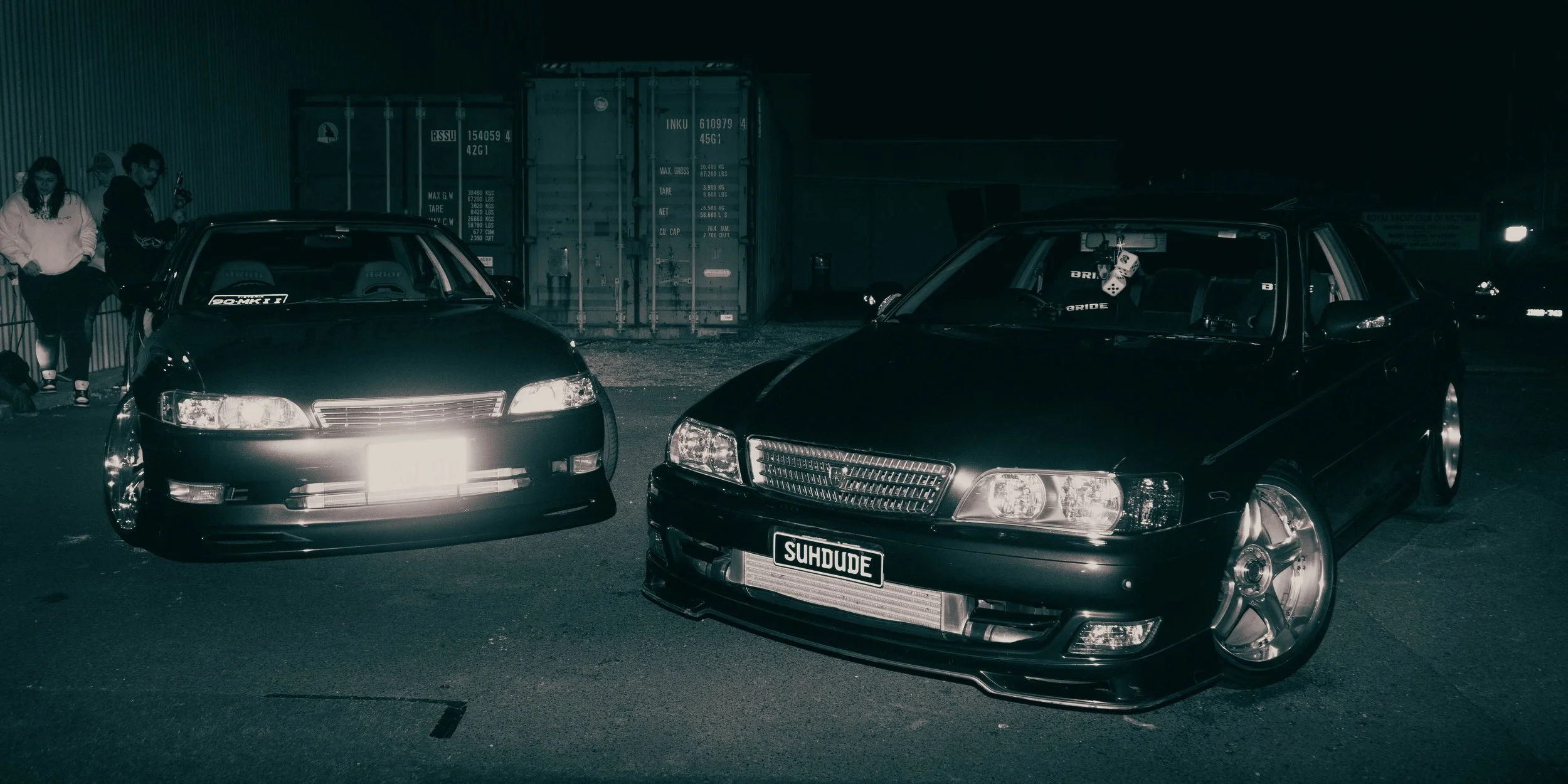 Two black cars parked side by side at night with two people standing near the cars and shipping containers in the background.