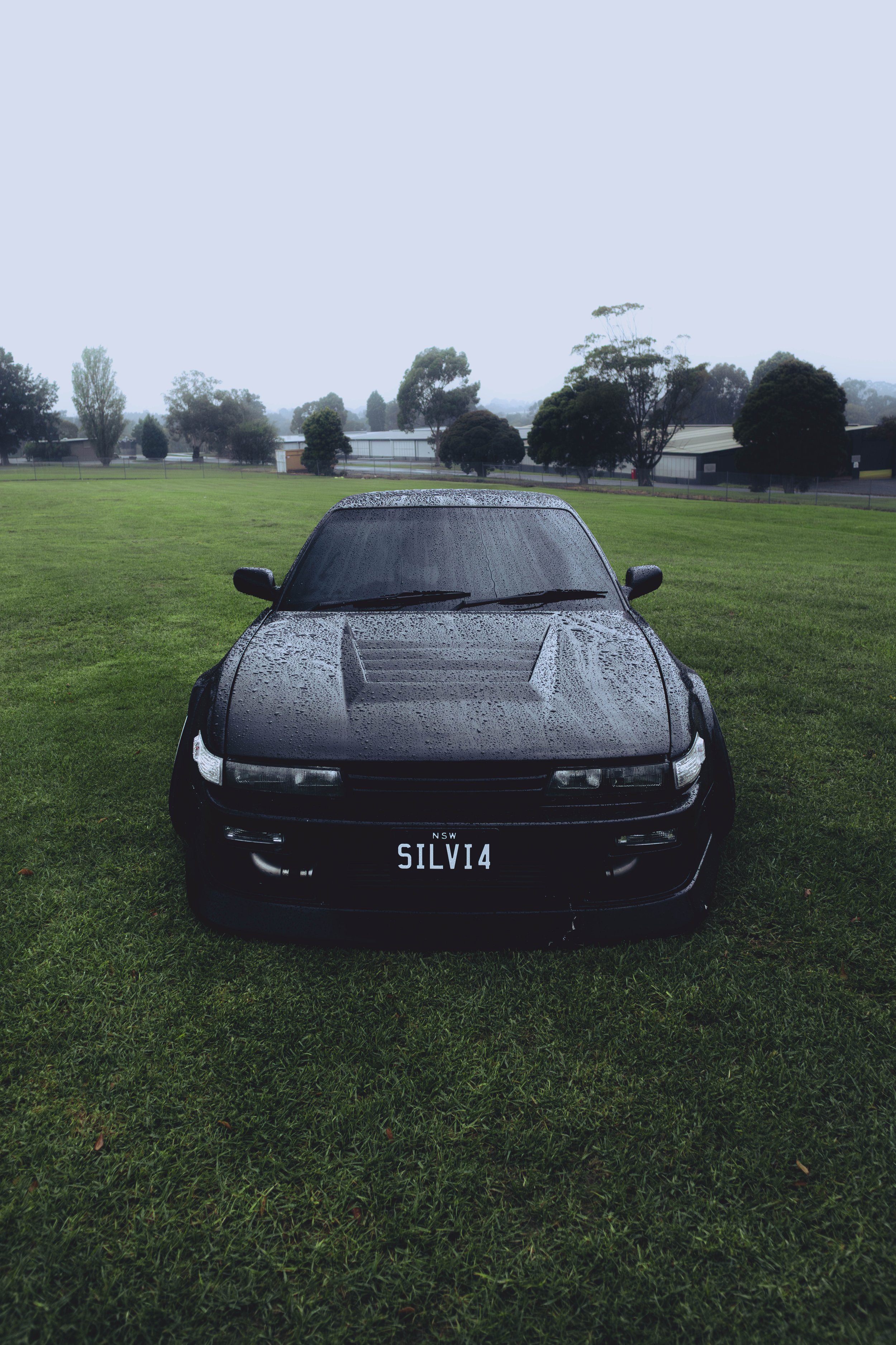 Black sports car with water droplets on the hood, parked on a grassy field on a rainy day, with trees and industrial buildings in the background.