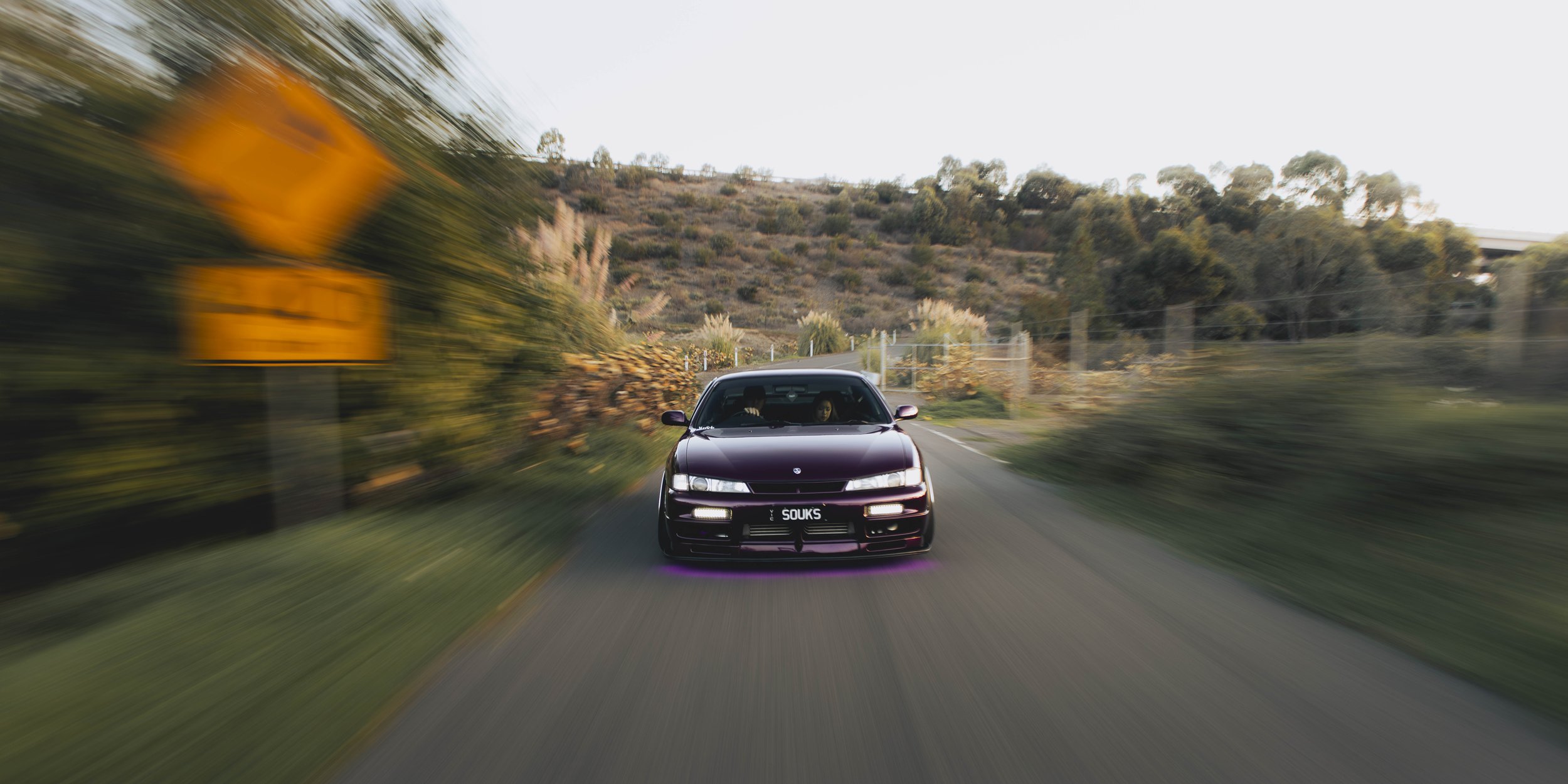 A purple Nissan S14 Silvia 200SX purple underglow driving on a rural road surrounded by trees and hills, with motion blur indicating speed.