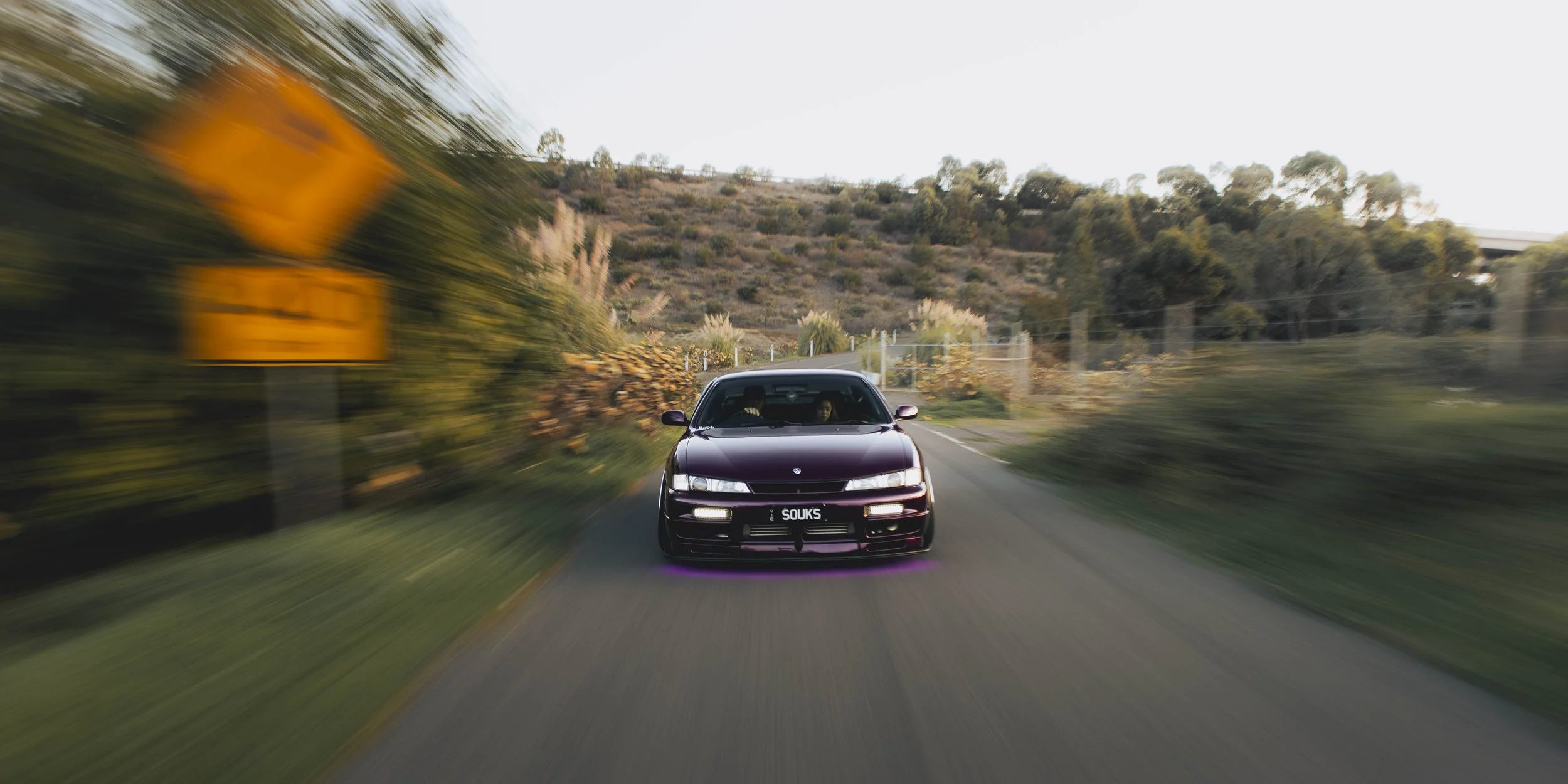 A purple Nissan S14 Silvia 200SX purple underglow driving on a rural road surrounded by trees and hills, with motion blur indicating speed.