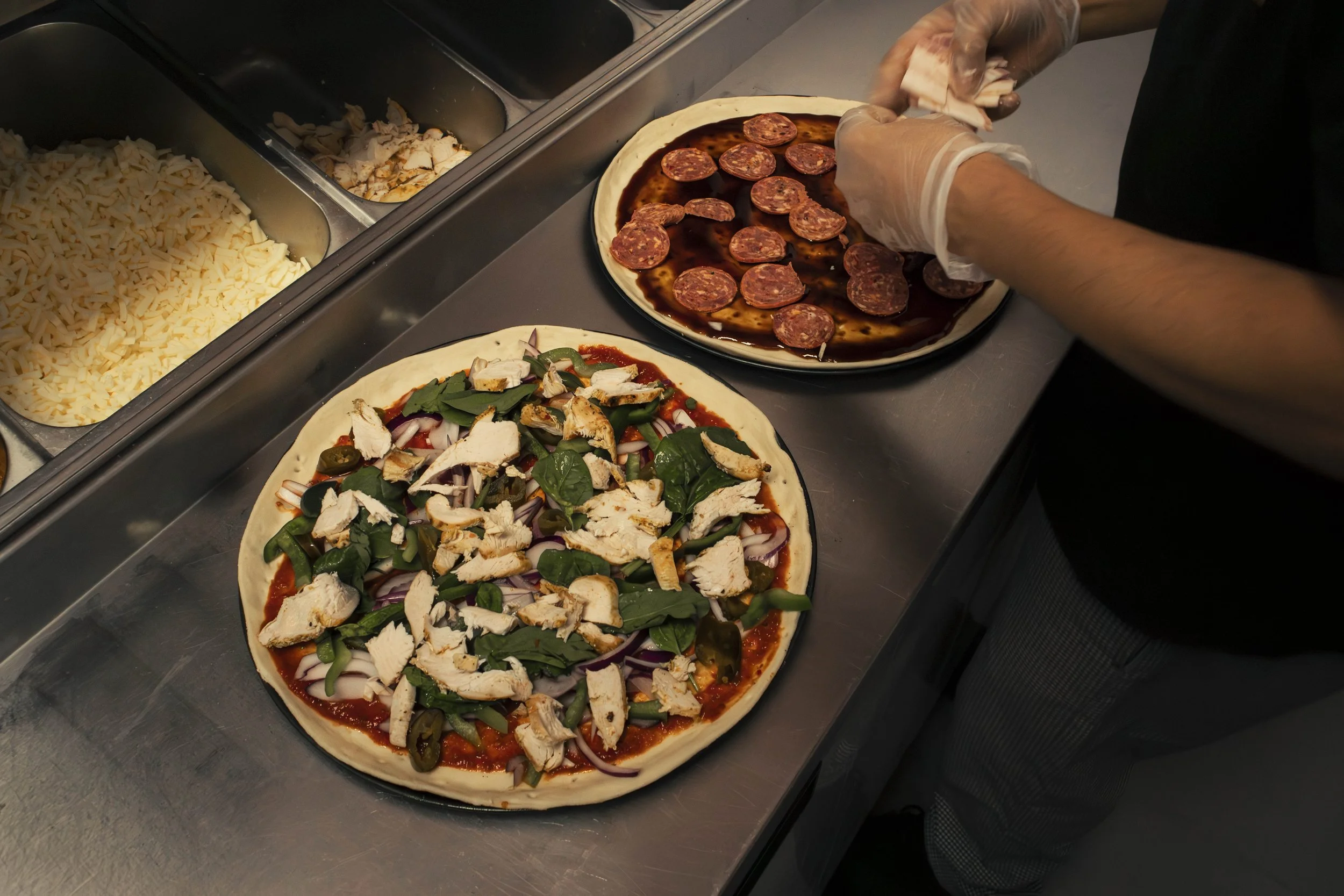 Person preparing a pizza with pepperoni slices on a tray, other pizza ingredients in metal containers nearby.