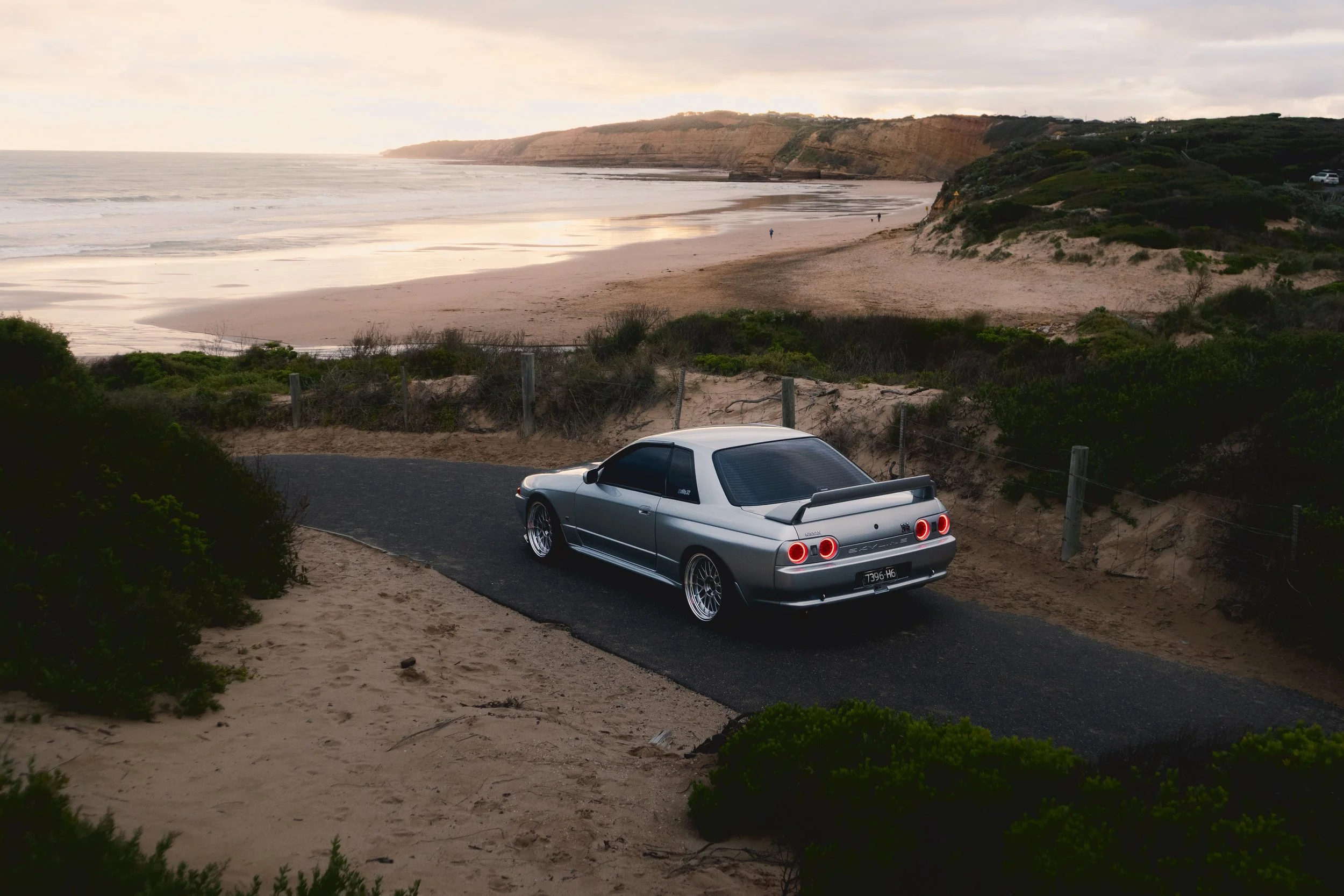 A silver sports car parked on a paved road overlooking a beach with cliffs in the background, under a cloudy sky.