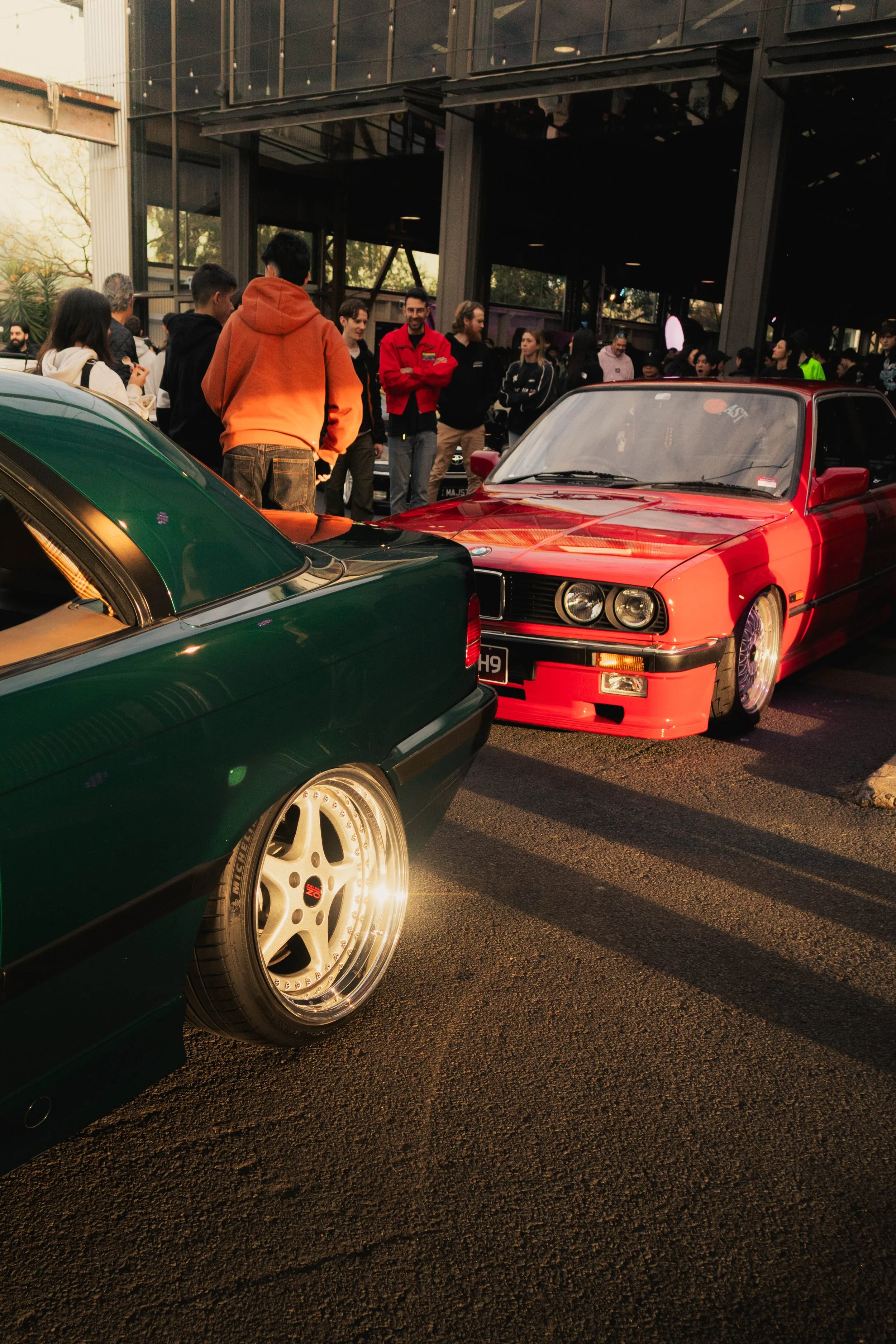 People gathered at a car show, looking at vintage and classic cars, including a red car in the foreground.