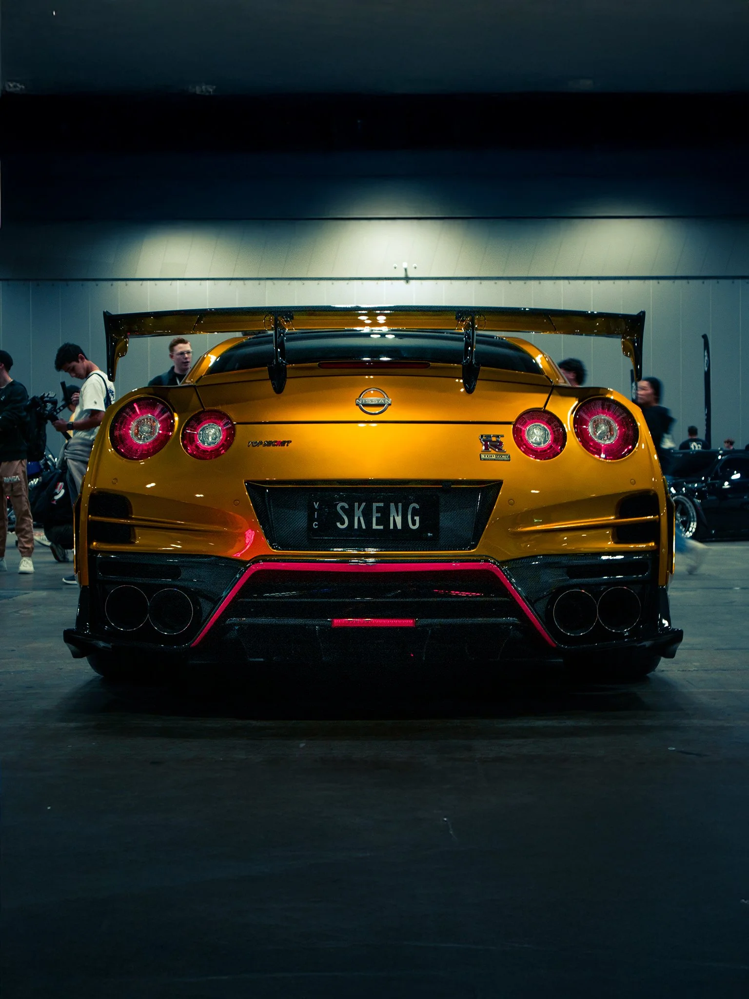 Rear view of a yellow Nissan GT-R sports car with a large rear wing, surrounded by people at an indoor car event.