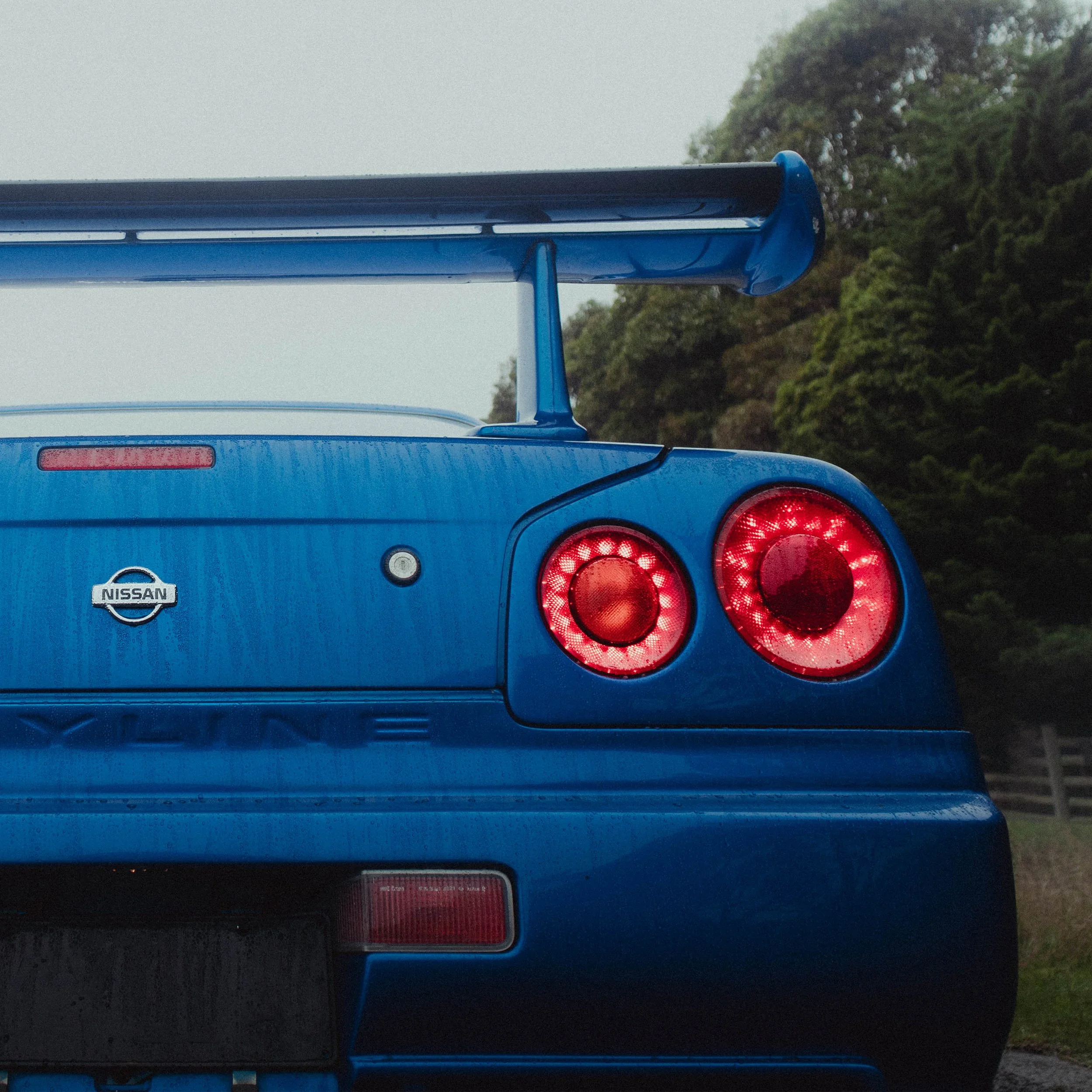 Rear view of a blue Nissan Skyline R34 showing taillights and a roof spoiler, with trees in the background.