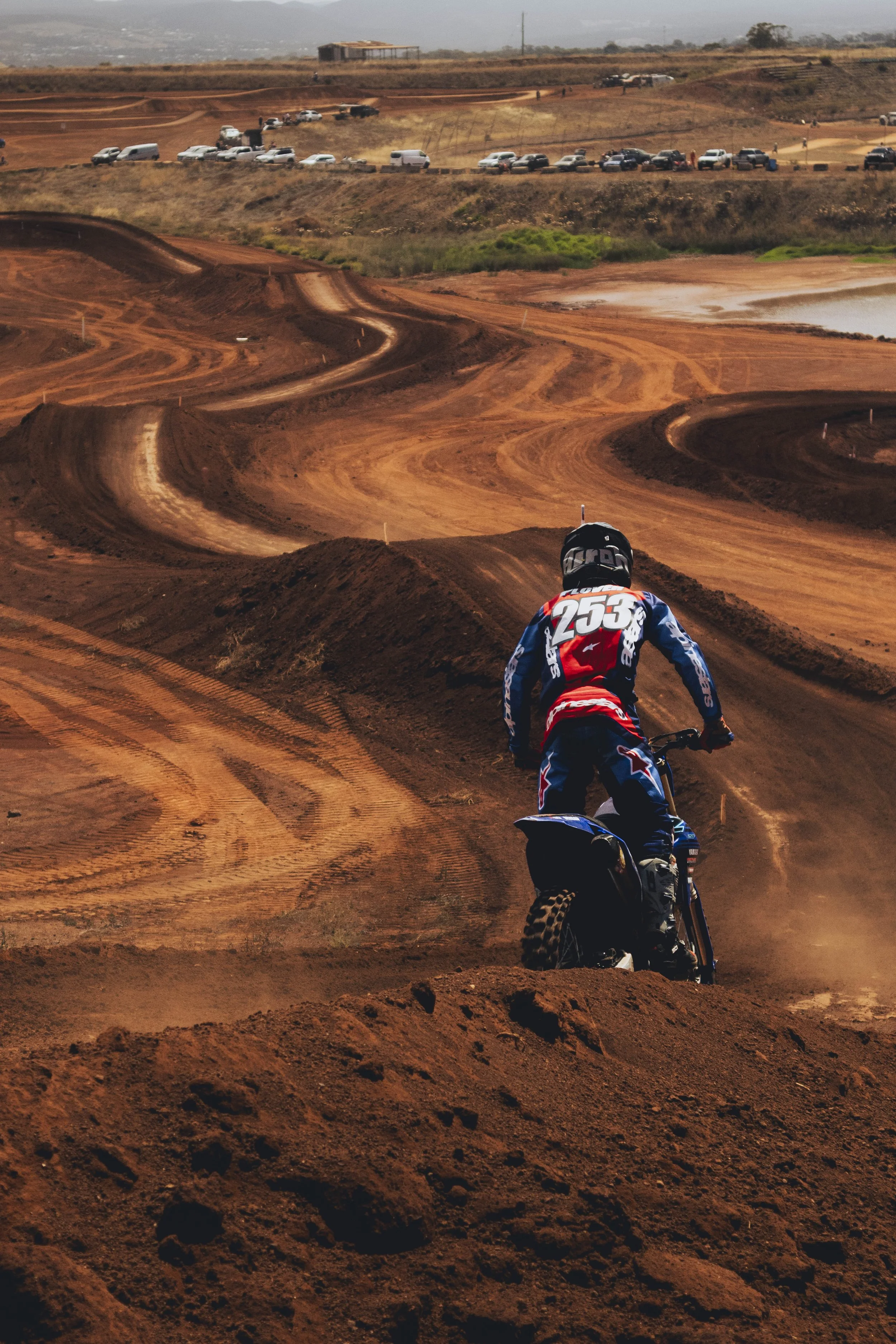A motocross rider, wearing a helmet and a blue, red, and white racing suit numbered 253, rides on a dirt track with sharp turns and mounds. The background shows several parked cars and a few small buildings on a hilly landscape under overcast skies.