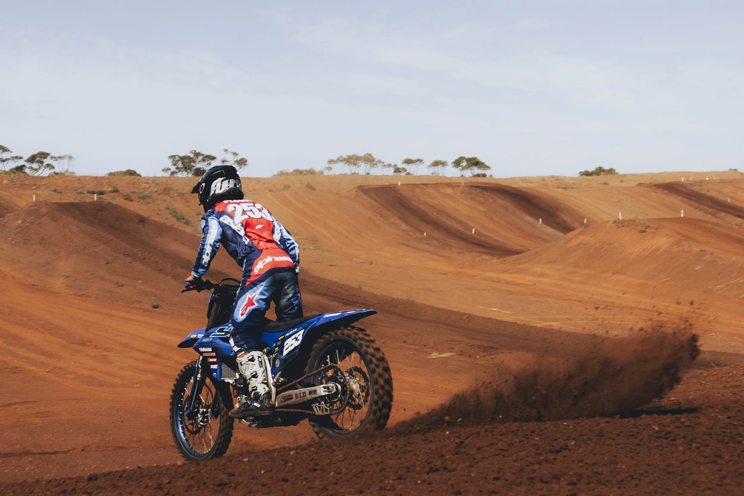 A motocross rider wearing a helmet and racing gear riding a blue dirt bike on a reddish-brown dirt track with rolling hills and scattered trees in the background.