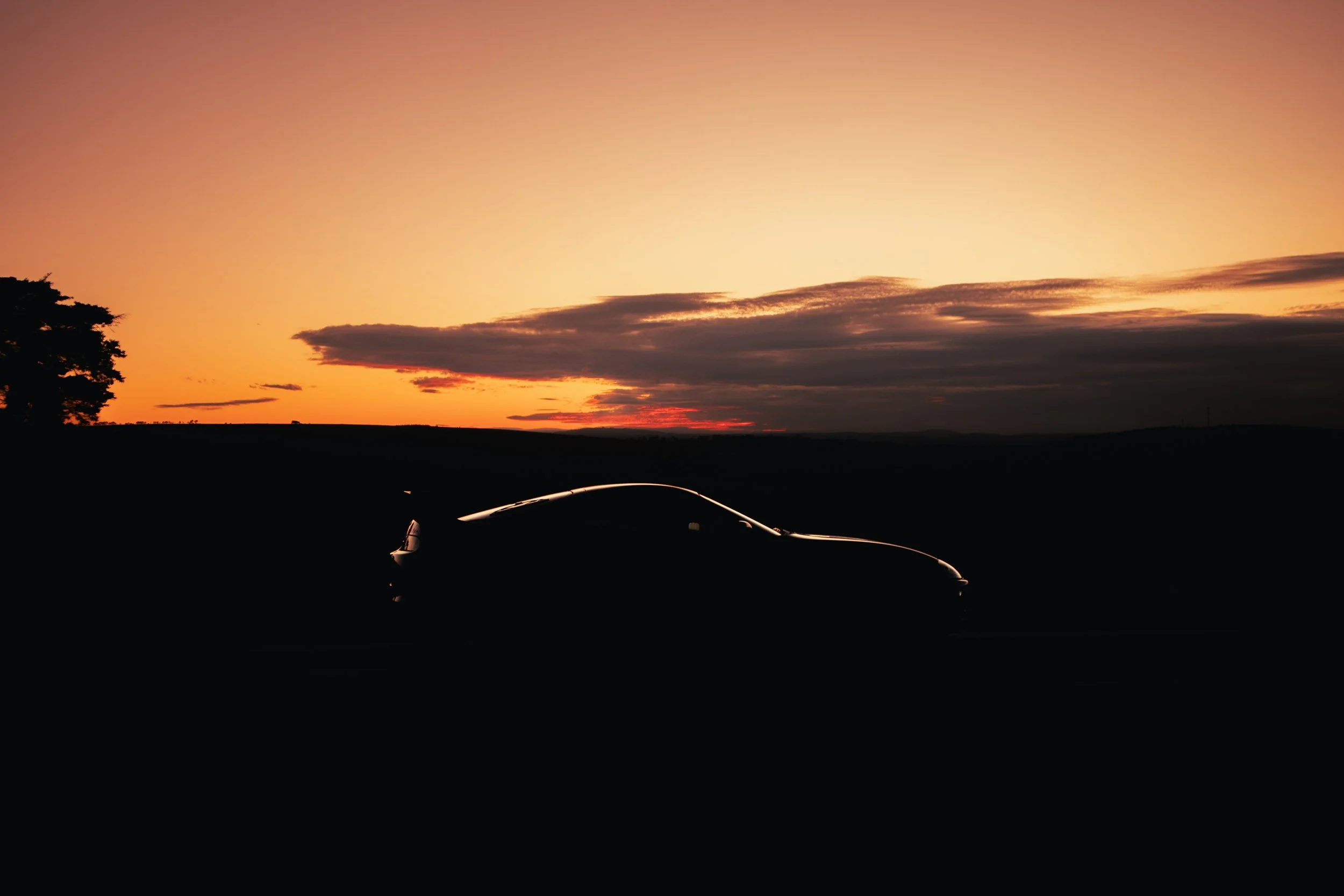 A car parked outdoors during sunset with a colorful sky, silhouetted trees, and clouds in the background.