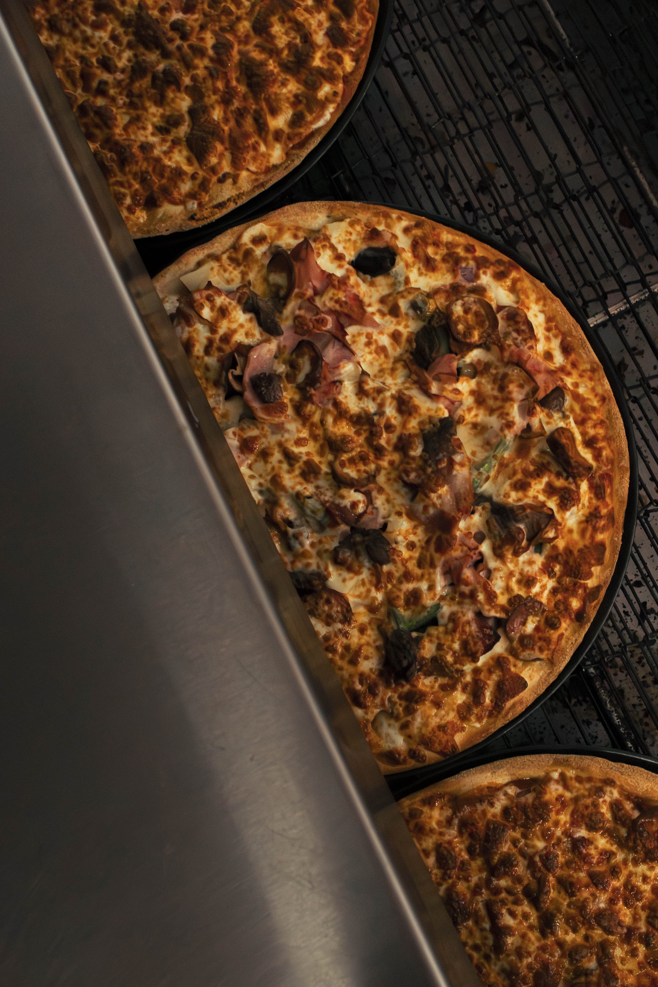 Three pizzas baking in an oven, with one pizza displaying toppings like mushrooms, ham, and green peppers, while another is topped with sausage and pepperoni. The pizzas are on a metal rack in the oven.