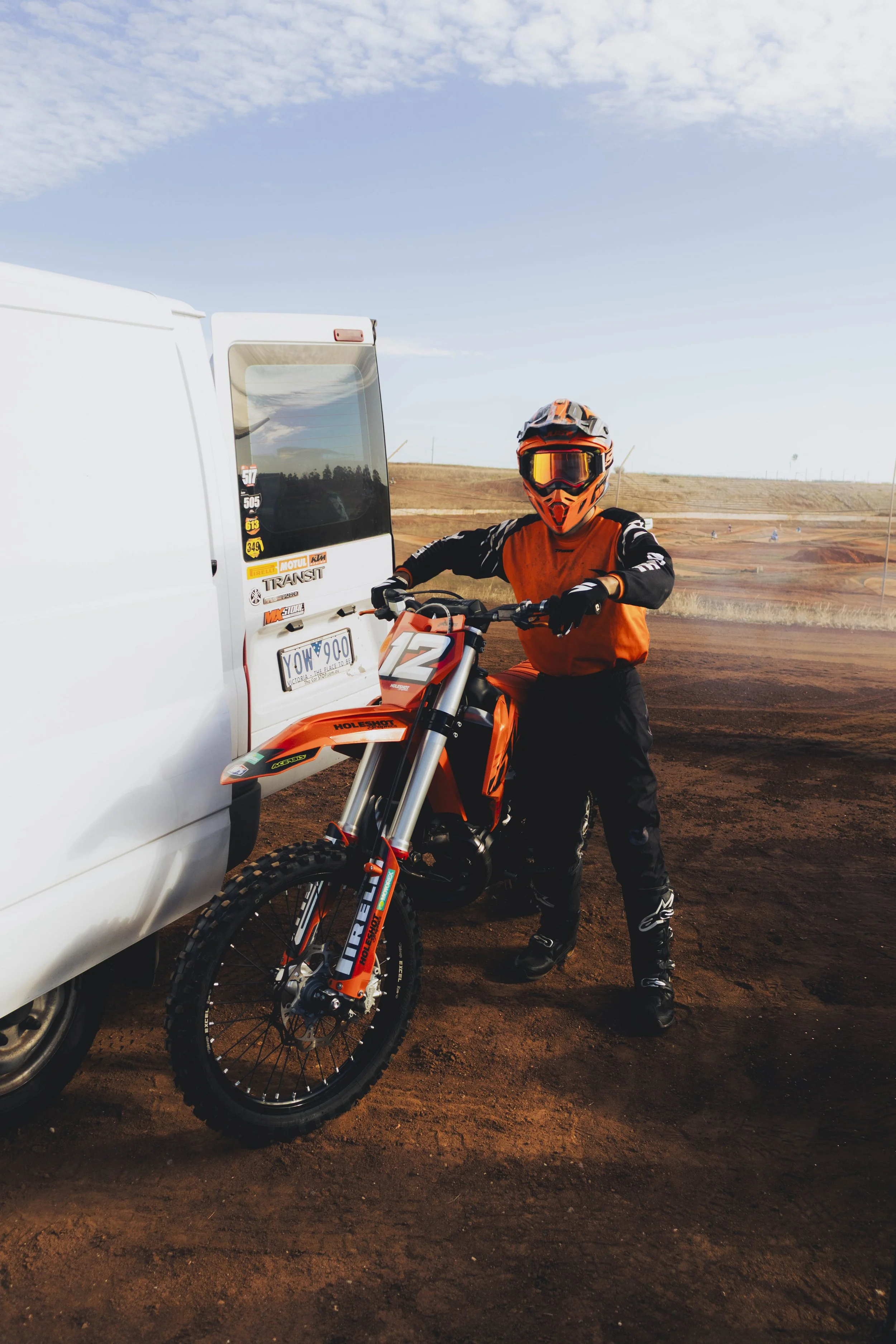 A motocross rider in orange gear and helmet sitting on an orange dirt bike, standing next to a white van with dirt track in the background on a clear day.
