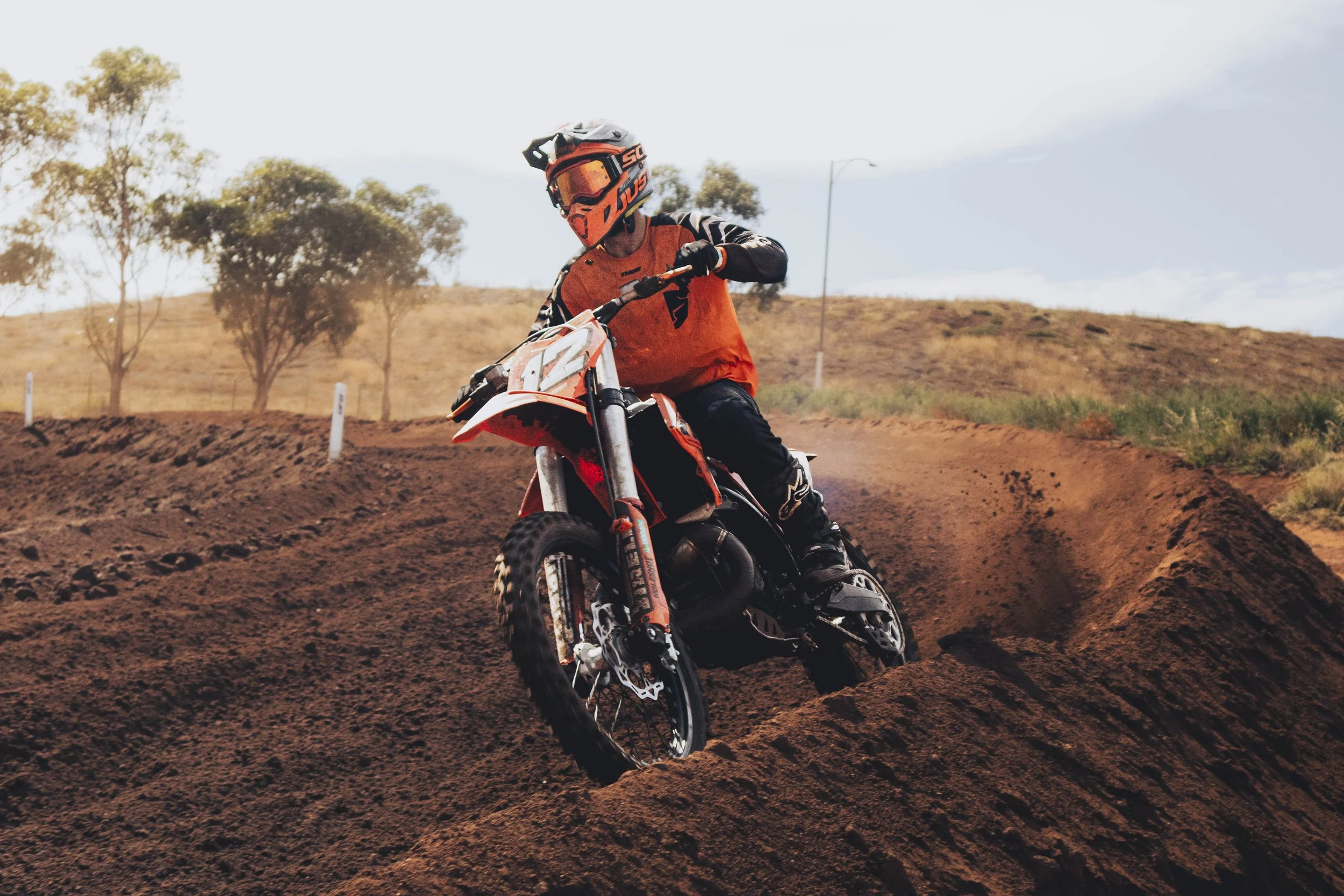 A person wearing an orange and black helmet and riding a dirt bike on a dirt track with trees and hills in the background.