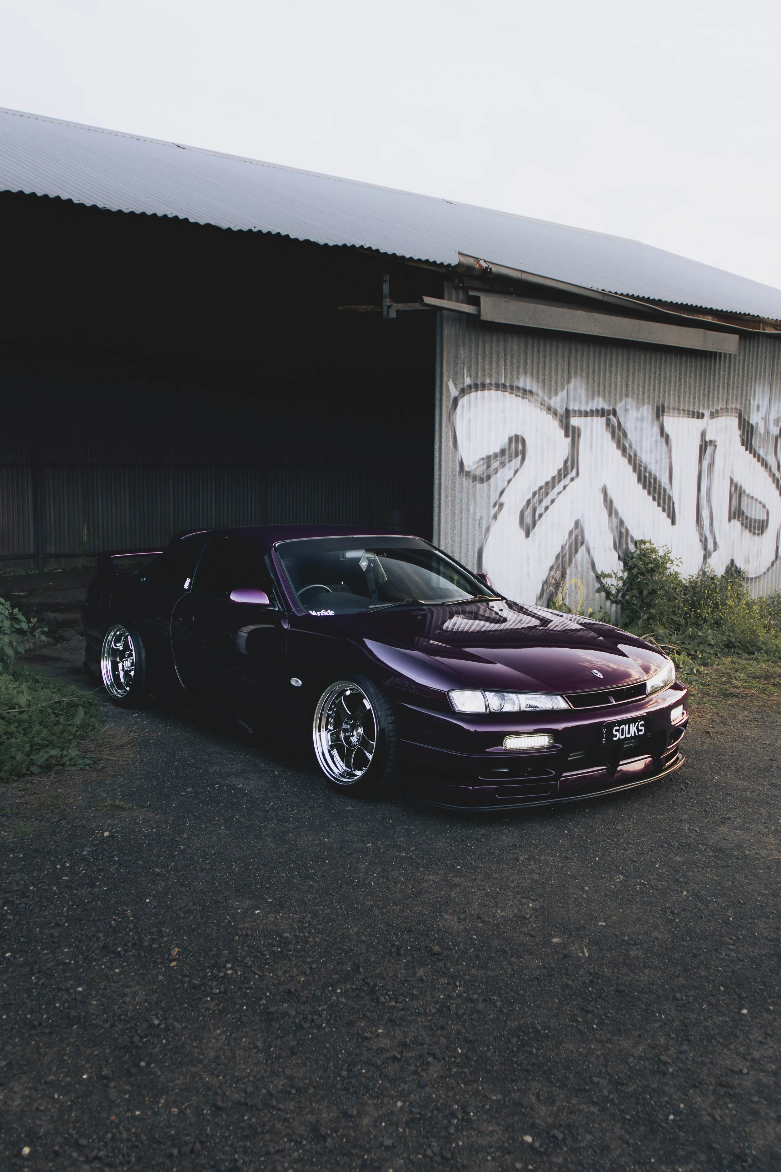 A dark purple sports car with tinted windows and custom wheels parked in front of a graffiti-covered metal warehouse with a corrugated roof.