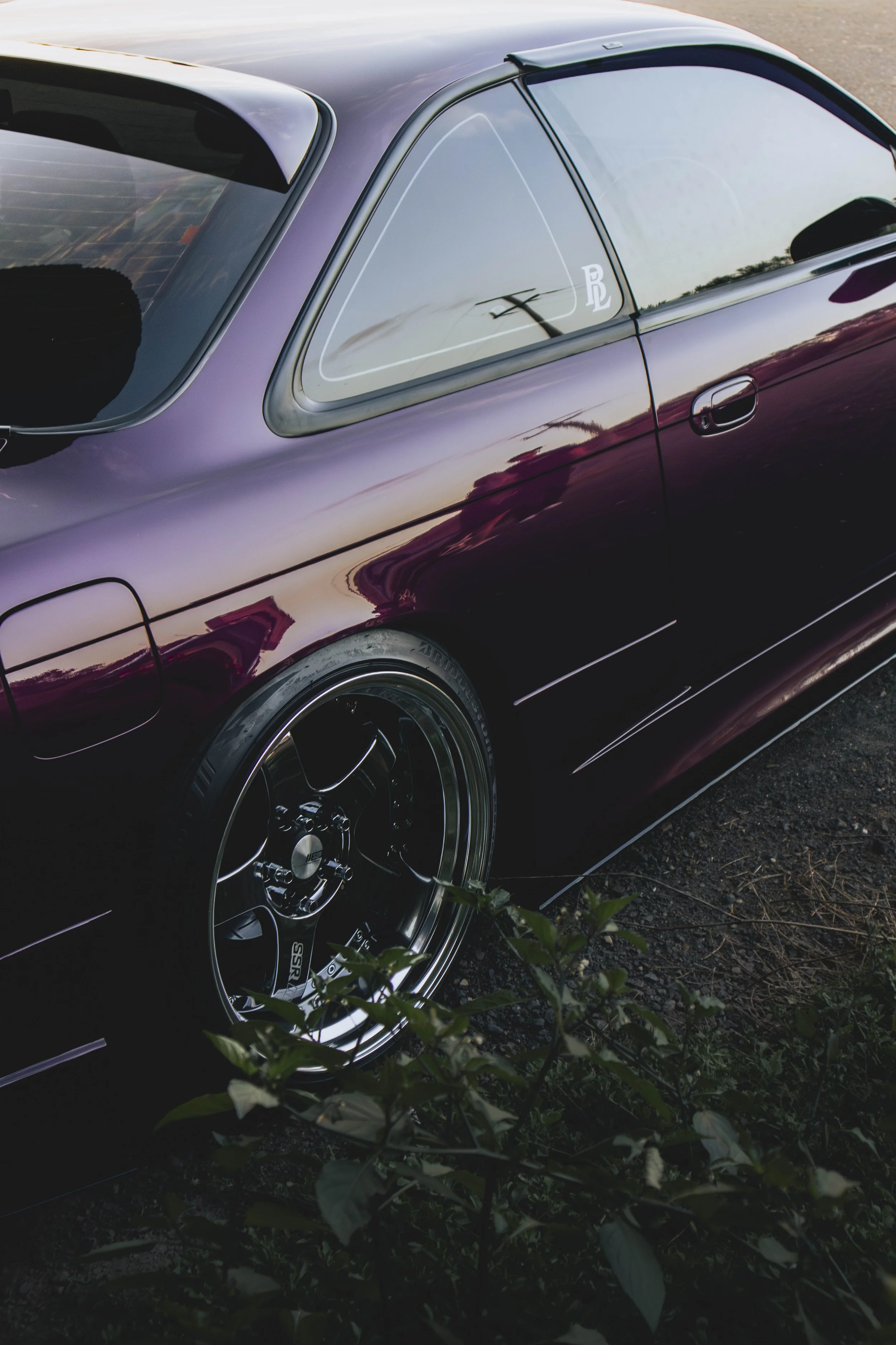 A purple sports car parked on a gravel surface with some green plants in the foreground, reflected sunset sky, and a parking lot in the background