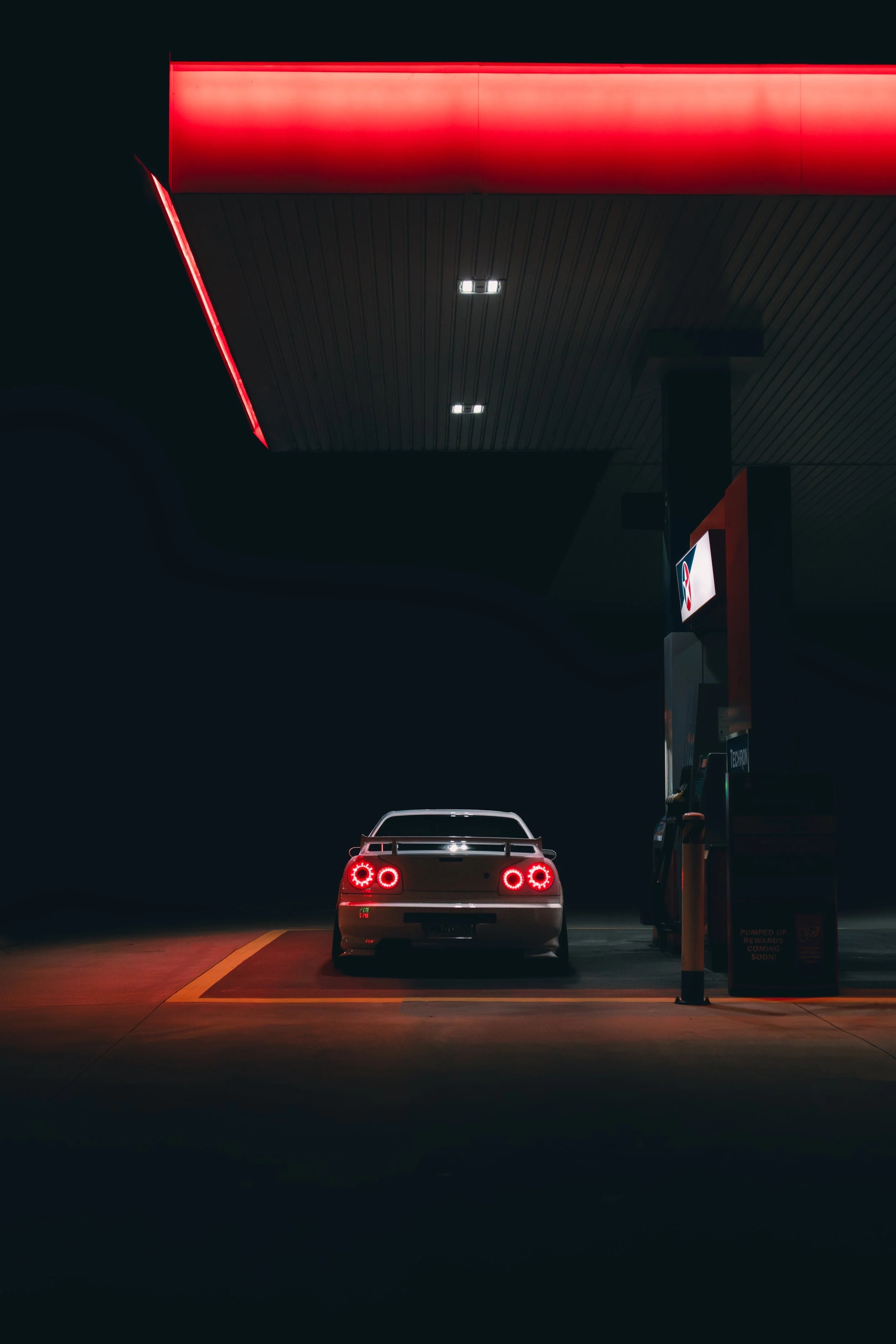 White Nissan Skyline R34 GTR parked at a gas station during nighttime, illuminated by overhead lights and red neon signs, with the car's rear lights glowing red.