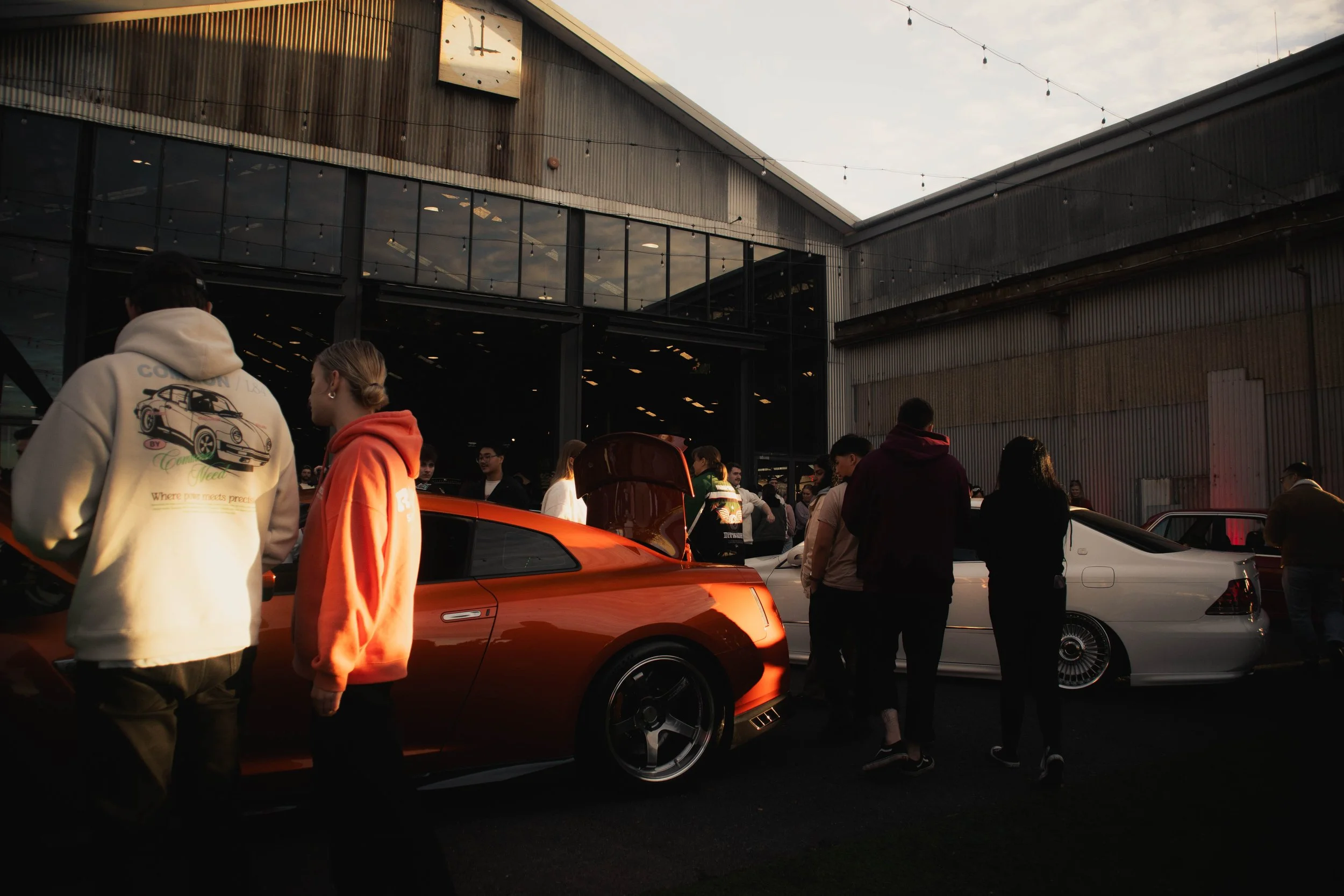 A group of people gathered at a car show outside a building with large glass windows and a metal exterior, with several sports cars, including an orange one in the foreground.