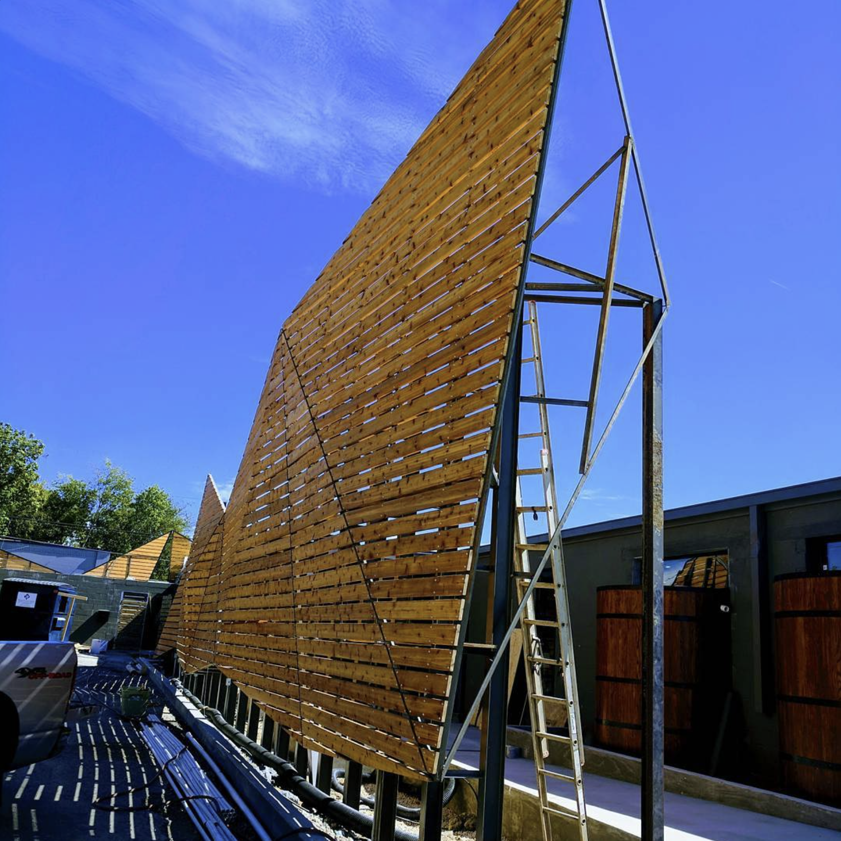 A large wooden structure under construction, with ladder and metal framework, in an outdoor setting with blue sky and surrounding buildings.