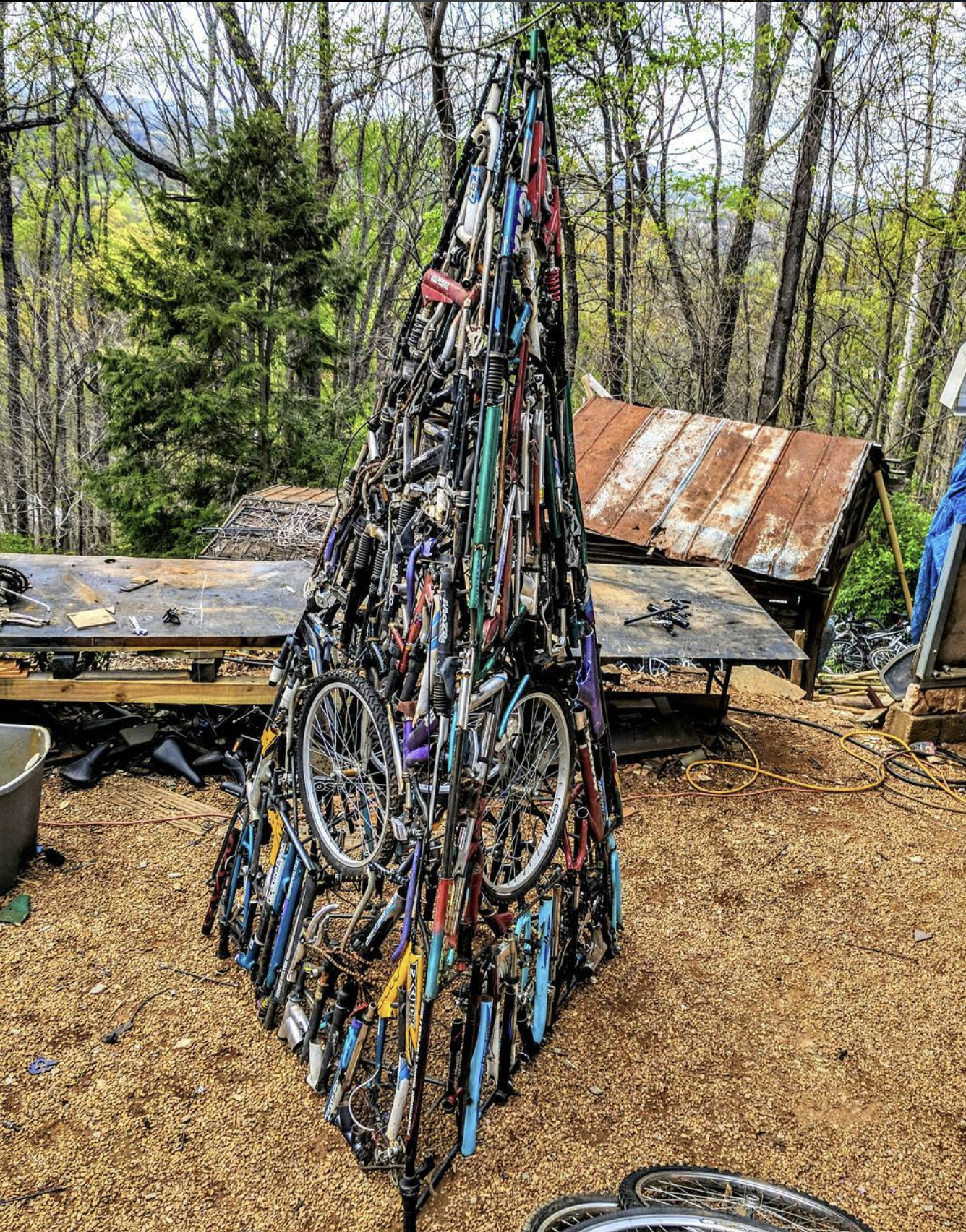 A sculpture of a mountain made from assembled bicycle parts, set outdoors on a dirt surface with trees and a rusty shed in the background.