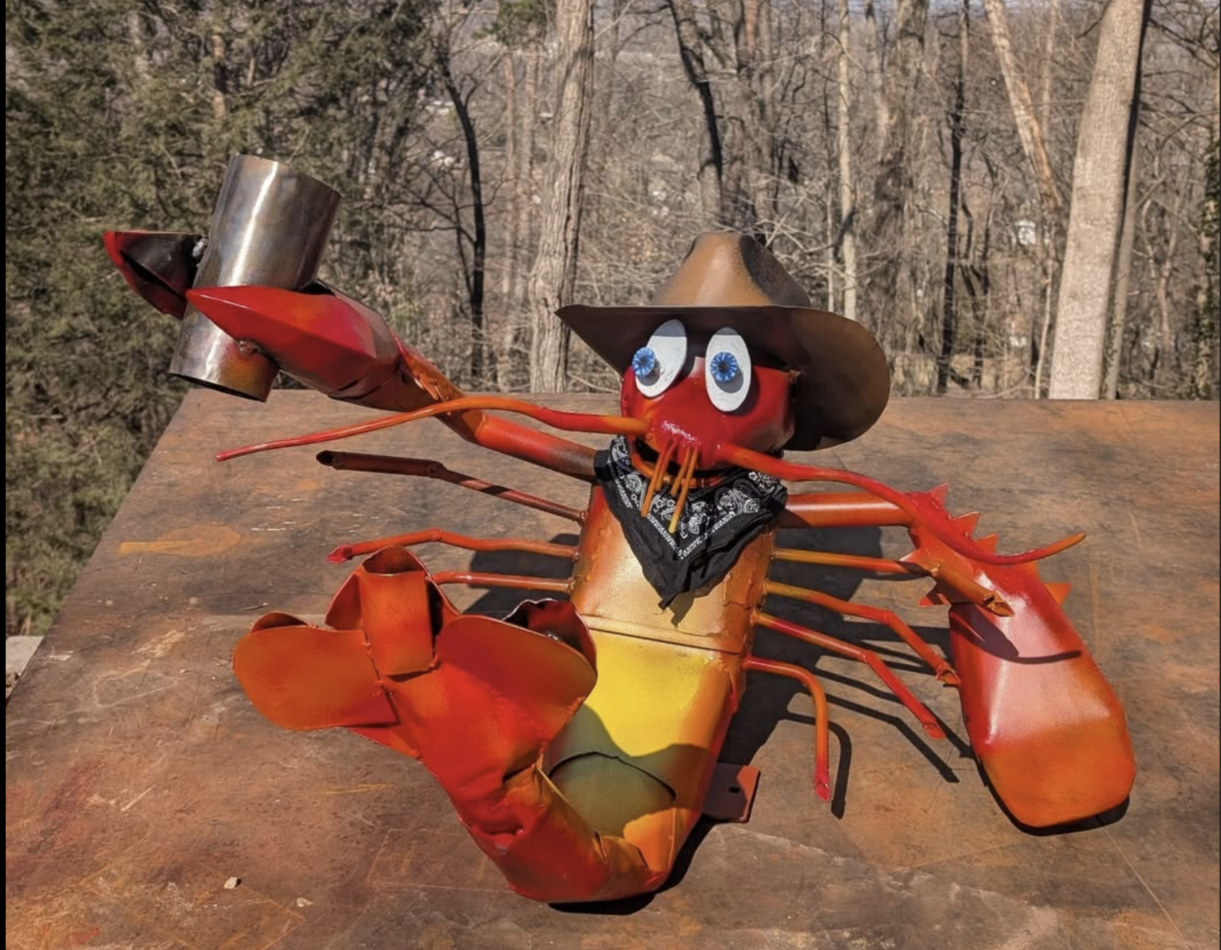 A whimsical metal lobster sculpture with cartoonish eyes, a cowboy hat, a bandana, and exaggerated claws, set outdoors against a wooded background.