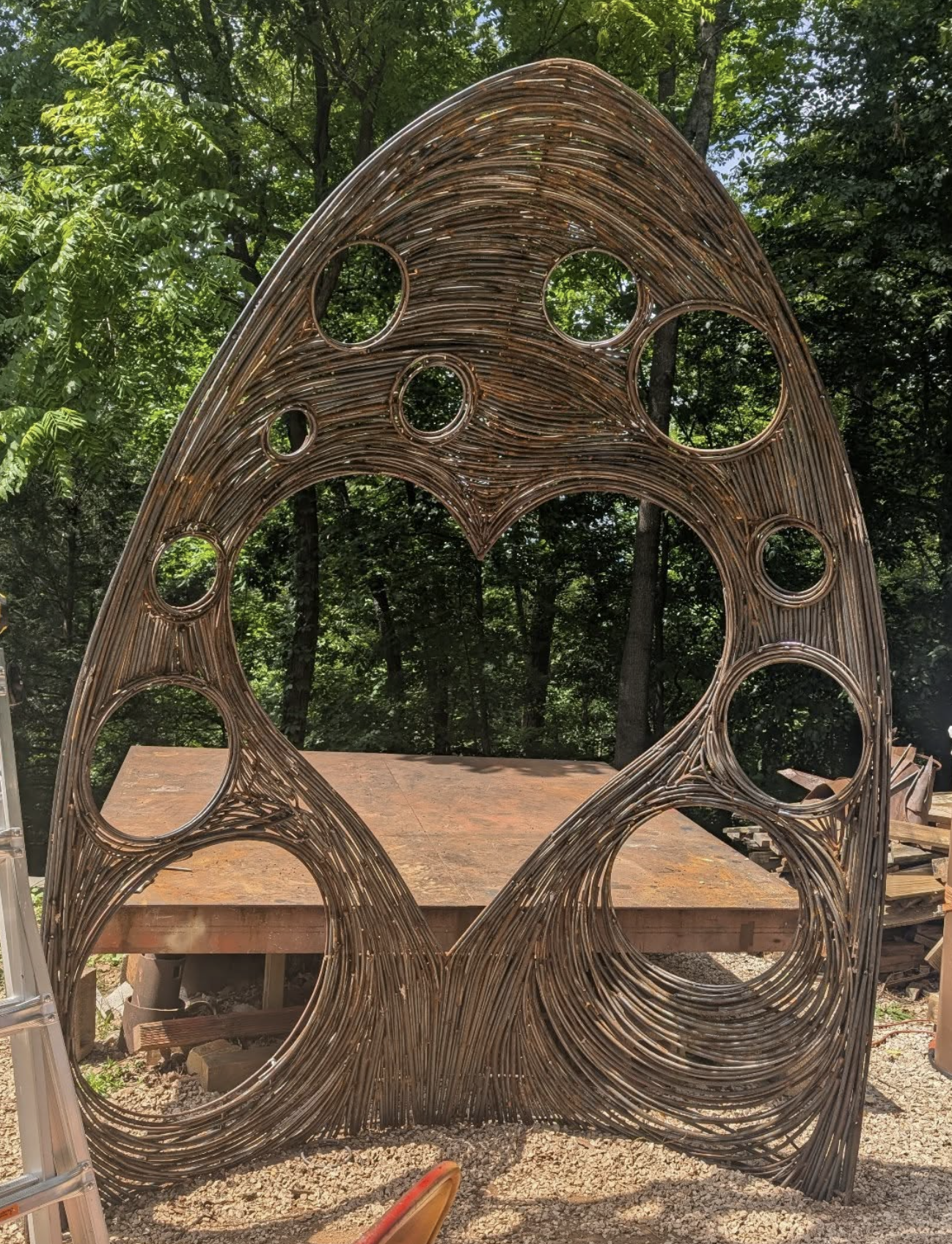 A large outdoor sculpture made of intertwined metal rods resembling a whimsical, abstract owl with multiple circular cutouts, set against a leafy green forest background.