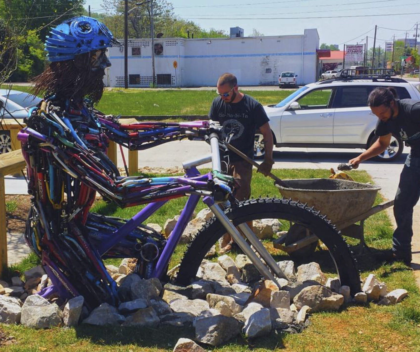 Two men working on a colorful sculpture of a human figure on a rock bed in an outdoor area, with cars and a white building in the background.