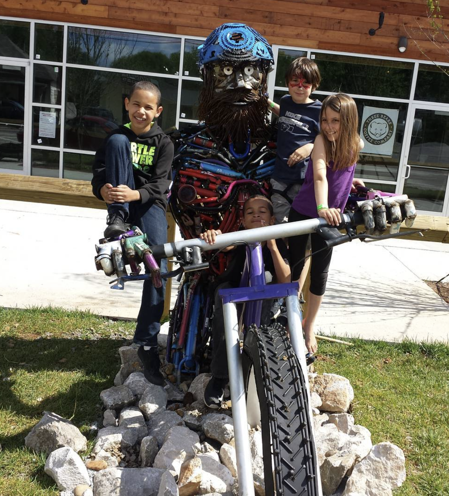 Group of five children posing with a large sculpture of a pirate, made from bike parts, outside a building with large windows.